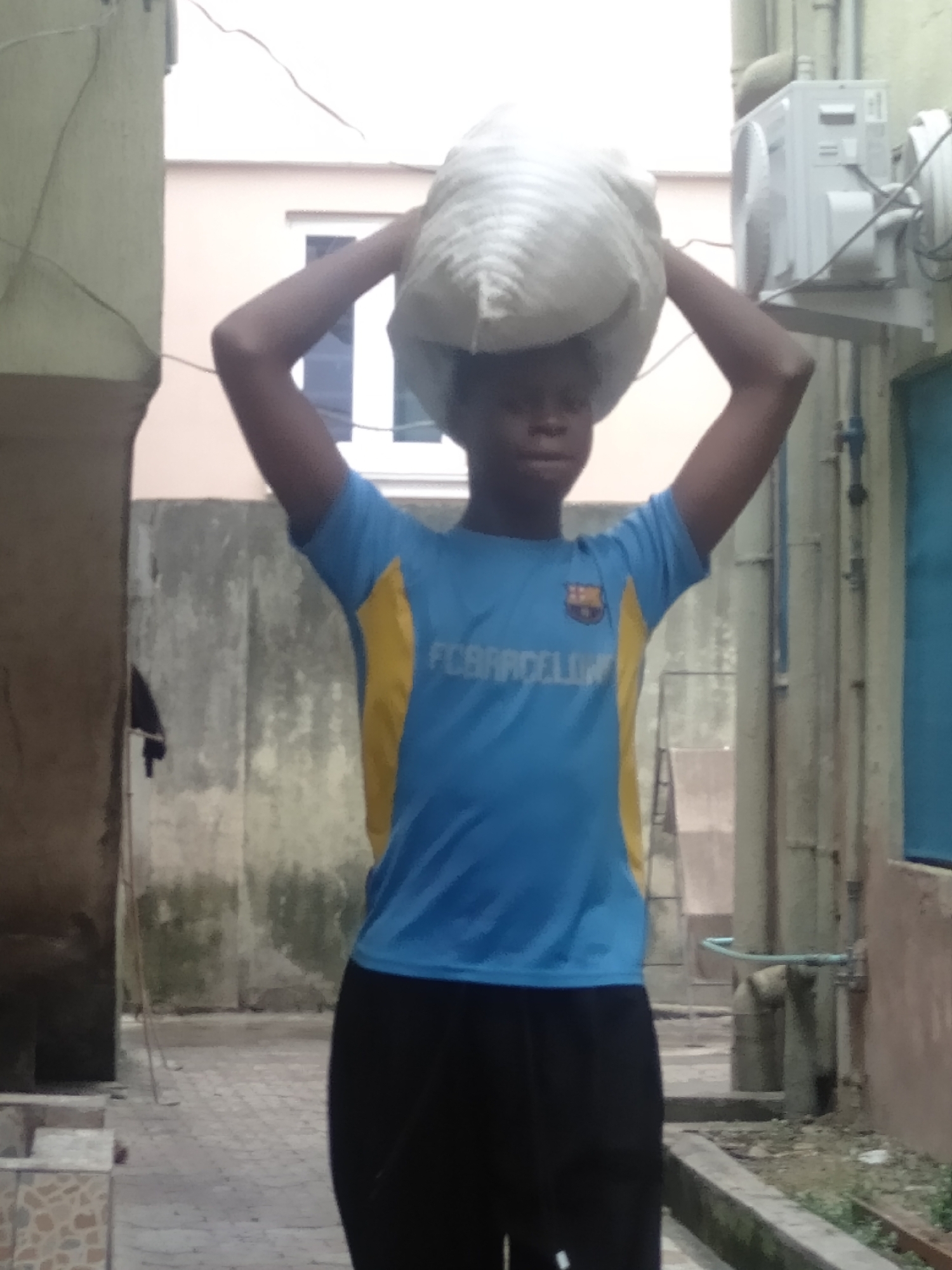 A young person, likely a boy, stands centered in an outdoor alleyway between buildings, balancing a large, light-colored sack or bag on their head. Their arms are raised, hands near the bag, seemingly to steady the load. They are wearing a blue and yellow short-sleeved t-shirt with "FC BARCELONA" and the club's emblem printed on the front, paired with black pants. The person's expression is neutral as they look forward.
The background shows weathered concrete or stucco walls. On the right, an outdoor air conditioning unit is mounted on a wall, accompanied by various pipes and wires. Further back on the right, a faucet with a blue hose is visible. The walls display areas of discoloration and moss, while some sections of buildings in the background are painted light beige or off-white, with a window visible in one. The ground is paved with light-colored, possibly interlocking, stones or tiles. In the bottom left corner, patterned tiled steps or a low wall can be seen.
The scene is illuminated by bright, diffused daylight, suggesting either clear or overcast weather conditions. The setting appears to be a residential or urban environment, possibly in a warm climate, with utilities like external air conditioning units and exposed plumbing visible. The activity depicted is the manual transport of goods using head-carrying.