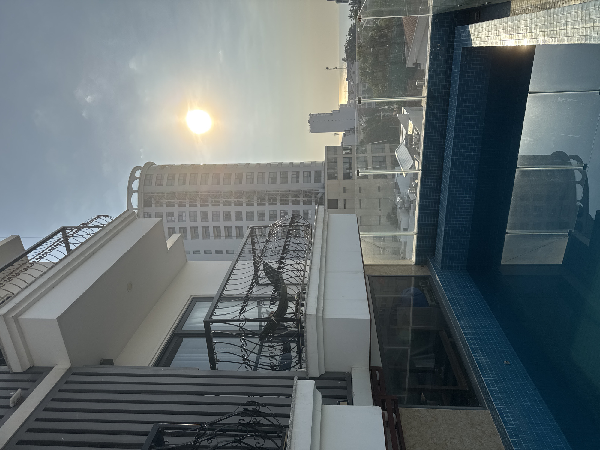 This image captures a rooftop view of Nha Trang, Vietnam, featuring a private swimming pool in the foreground and a city skyline in the background, bathed in the warm glow of the setting sun. The pool is tiled with small, dark blue squares and reflects the sky and the surrounding buildings. A glass fence encloses the pool area, offering an unobstructed view of the cityscape.
Dominating the mid-ground are several tall buildings, characteristic of a coastal city. One particularly prominent building is a cylindrical structure with a tiered top, featuring numerous windows. To its right, a slender, rectangular skyscraper stands tall. Further in the distance, the ocean is visible, meeting the horizon under a bright, sun-drenched sky.
In the foreground, to the left, is a section of a building with a balcony. The balcony is adorned with ornate black metal railings and has a hammock suspended from it. Vertical gray slats form a decorative element on the side of this building. The overall scene suggests a luxurious resort or private residence, offering a serene and picturesque view of Nha Trang.