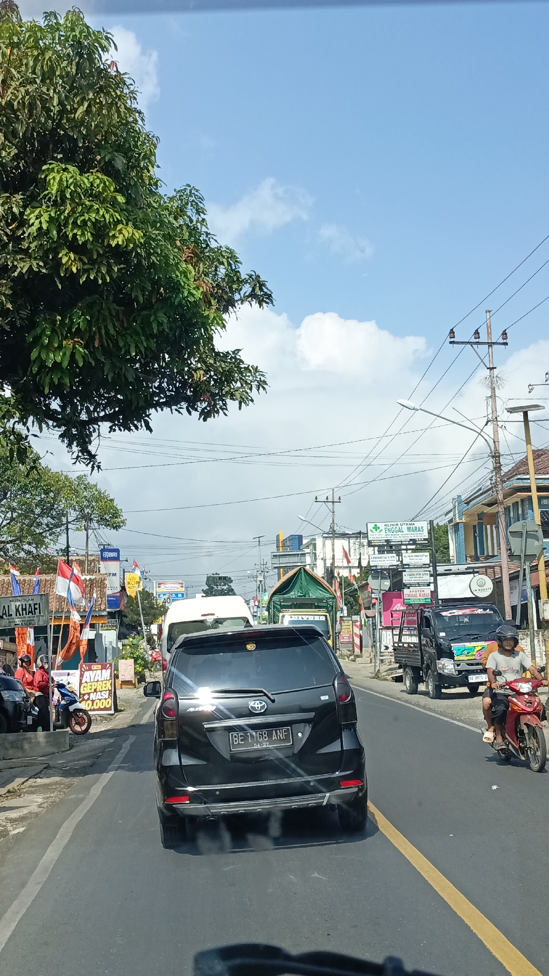 The image captures a street scene in Sukabanjar, Indonesia, likely during daytime given the bright sunlight and blue sky with scattered clouds. The perspective is from inside a vehicle, looking forward. A black Toyota Avanza is the prominent subject in the foreground, with its rear license plate reading "BE 1168 ANF". The vehicle is on a paved road with a yellow line marking the lane.
To the left of the road, there are various shops and stalls. One sign reads "MUSHOLLAH AL KHAFI" and another displays "AYAM GEPREK NASI 10.000". Red and white Indonesian flags are visible. A motorcycle is parked near these establishments, and two individuals in red uniforms are standing nearby.
The road is moderately busy with traffic. A white van is ahead of the Avanza, and further down the road, several trucks are visible, including a black truck with colorful graphics. A person on a red motorcycle is riding in the right lane, to the right of the black Avanza.
On the right side of the street, a clinic named "KLINIK UTAMA ENGGAL WARAS" is advertised with a green sign. Utility poles with power lines run along the side of the road. Buildings with various architectural styles line the street, suggesting a commercial or mixed-use area.
A large tree with dense green foliage dominates the upper left portion of the image, casting some shadows. The overall impression is a typical, active street scene in a Southeast Asian city.