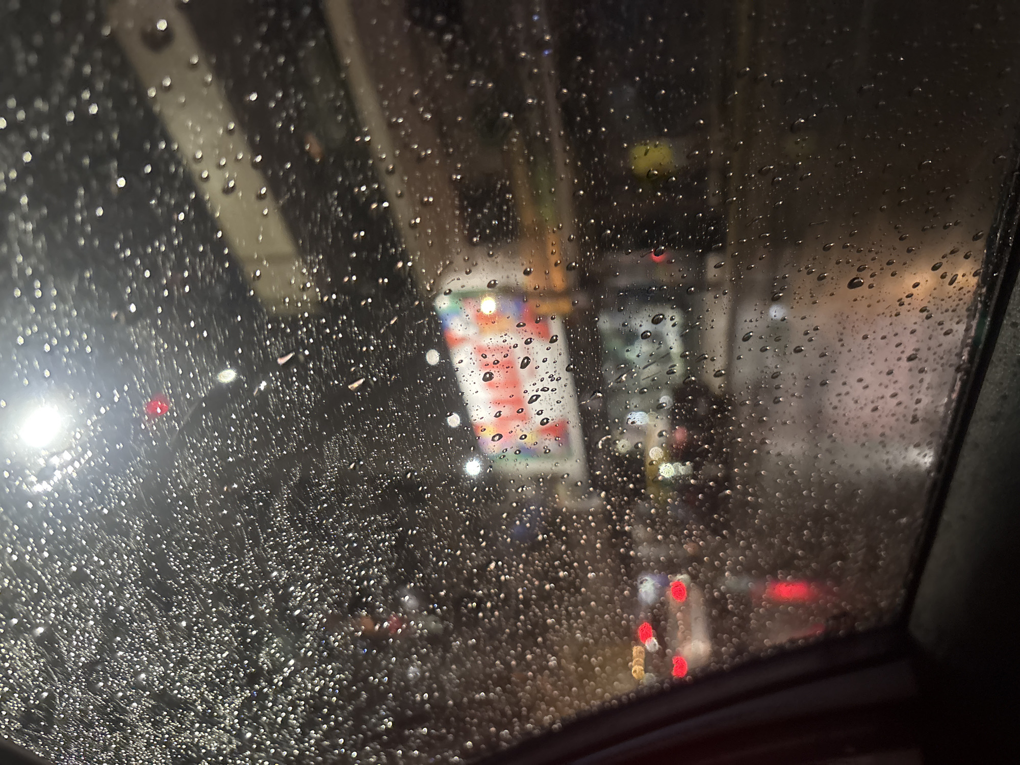 The image appears to be taken from inside a vehicle, likely a car, looking out through the rain-streaked windshield. Visible through the windshield are blurred city lights, traffic lights, and other vehicles. The rain obscures details, but suggests an urban setting at night. The overhead structure suggests a highway overpass. The location is Buenos Aires, Argentina, but specific geographical details are not discernible from the image alone. No people are visible. The focus is on the rain-covered windshield and the obscured city view.