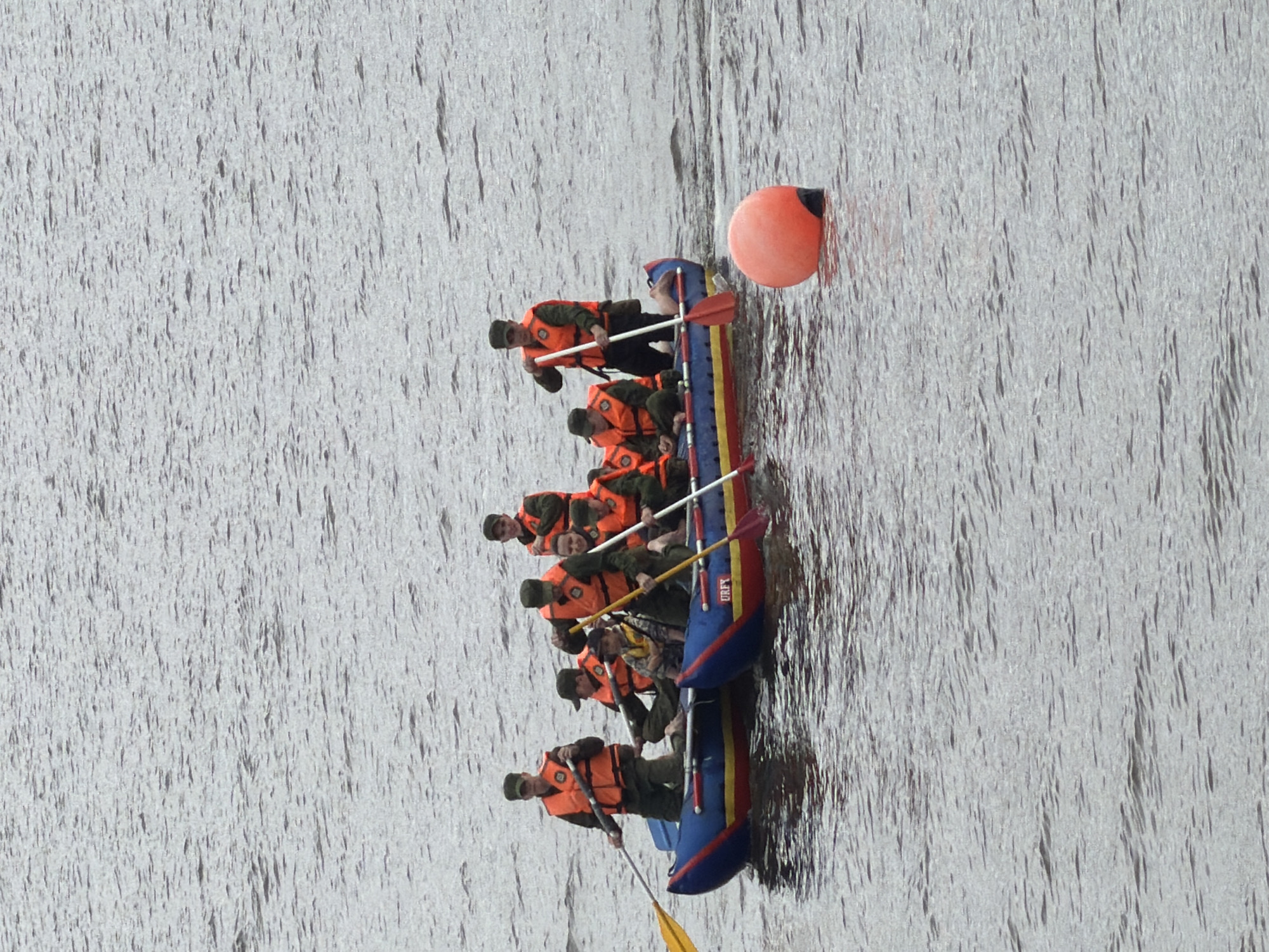 In Votkinsk, Russia, a group of eight individuals, all wearing orange life vests and military-style hats, are navigating a blue and yellow raft on a body of water. Seven individuals are actively paddling with oars, while one individual in the back is partially visible and not engaged in paddling. The water is choppy, with ripples and waves visible across the surface. In the foreground, a large, bright orange buoy with a black base is floating on the water. The overall scene suggests a training exercise or recreational activity on the water.