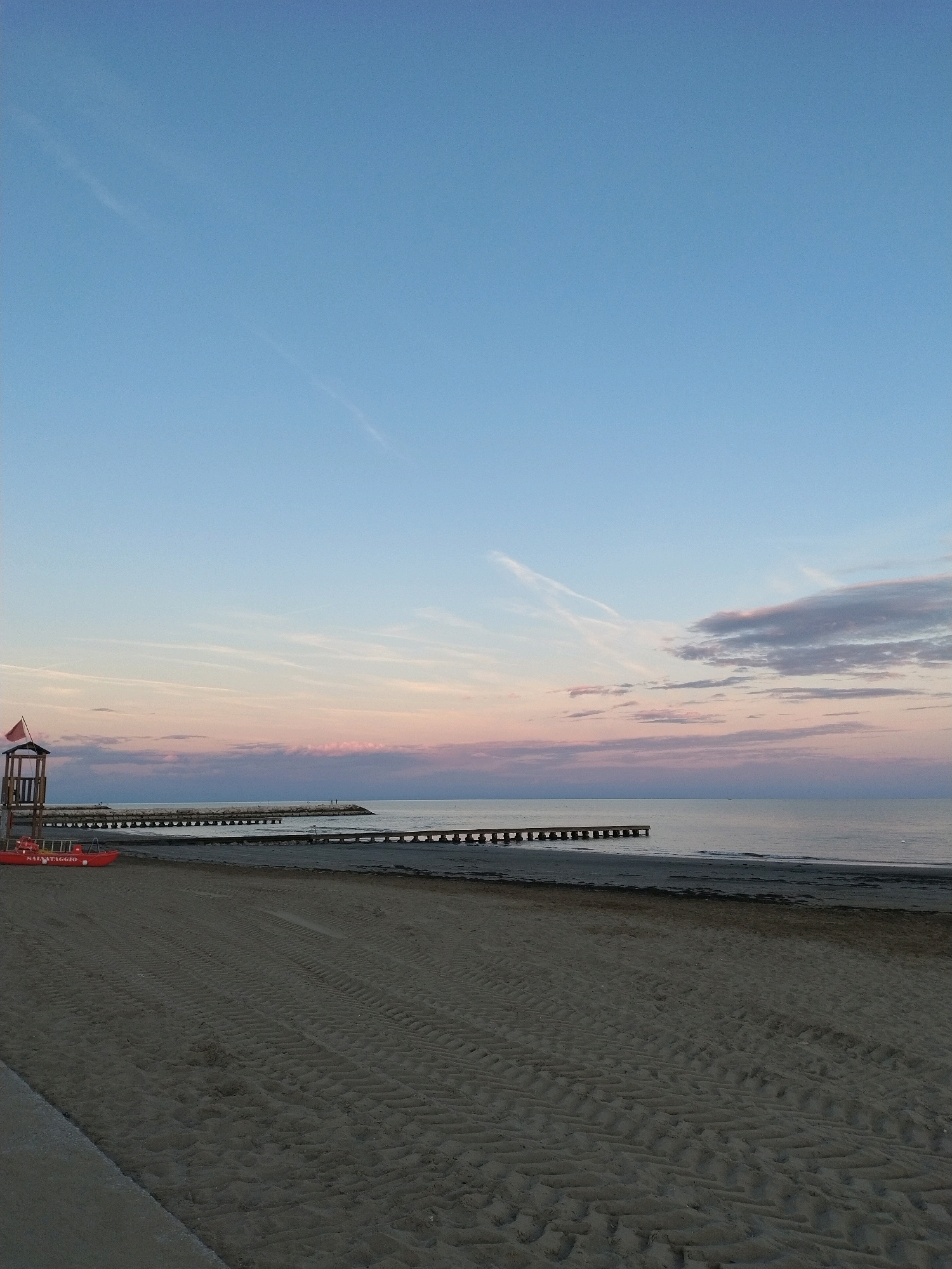 A beach scene in Caorle, Italy, captures the calm atmosphere of twilight. The sand in the foreground shows tire tracks, suggesting recent activity by a vehicle. On the left, a red lifeguard boat labeled "SALVATAGGIO" rests on the sand, near a wooden lifeguard tower topped with a red flag. The calm sea stretches to the horizon, where it meets a pastel sky transitioning from pale blue to soft pinks and grays from the setting sun. Several wooden jetties or piers extend into the water, spaced out along the coastline. The overall scene is serene and devoid of people, emphasizing the quiet beauty of the beach at dusk.