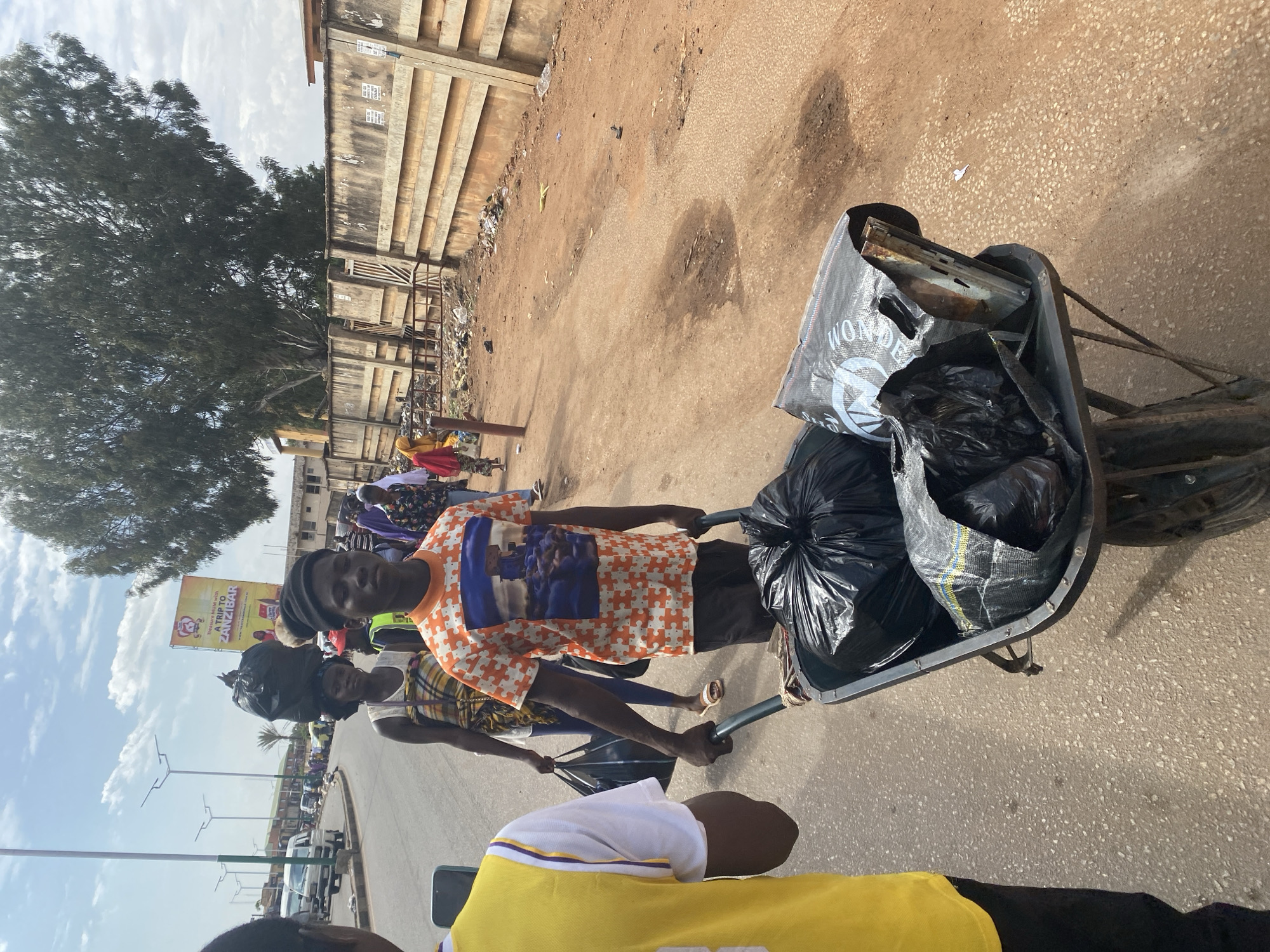 The image depicts a scene in Jos, Nigeria. A person in an orange and white puzzle piece patterned shirt pulls a wheelbarrow full of black trash bags and scrap metal on a paved road. Another person is behind them, slightly obscured. A third person in a yellow and white shirt is in the foreground. In the background are buildings and a large tree. Along the road, streetlights are visible. A sign advertising "A TRIP TO ZANZIBAR" can be seen.