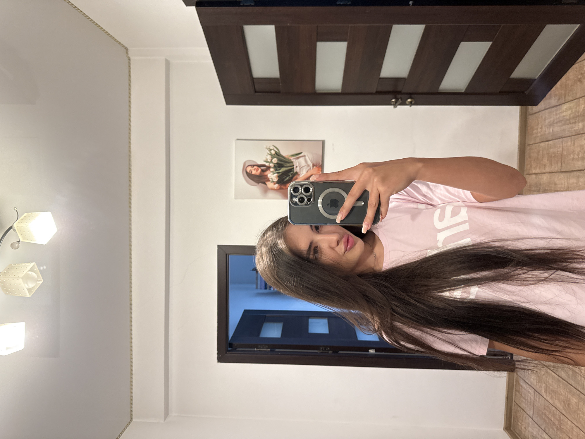 The image is a selfie taken by a young woman in a hallway. She is standing in front of a mirror, holding a smartphone to take the photo. She has long, dark brown hair and is wearing a pink t-shirt with white text. The hallway has white walls and a wooden floor. There are two doors visible in the background, one ajar to reveal a glimpse of another room, and the other a dark wood door with frosted glass panels. A framed picture of a woman holding a bouquet of flowers hangs on the wall. The lighting suggests an indoor setting, possibly a home or apartment. No specific details in the image allow for identification of a location within Chișinău, Moldova.