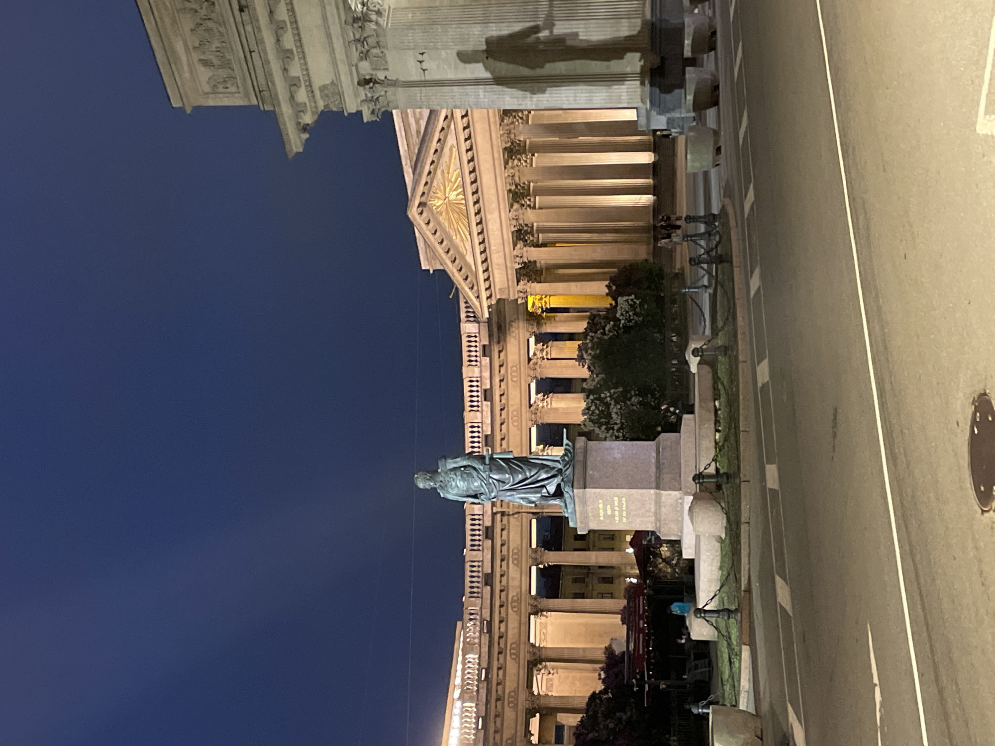 The image depicts a night view of Palace Square in Saint Petersburg, Russia. In the foreground is a bronze statue of Alexander I, positioned on a stone pedestal. The statue is surrounded by a low chain fence. The General Staff Building, a significant example of neoclassical architecture, serves as the backdrop to the statue. The building's facade is illuminated, revealing its colonnades, arches, and central triumphal arch topped with a quadriga sculpture. A street runs along the side of the square with street markings and a manhole cover. The sky above is a deep blue.