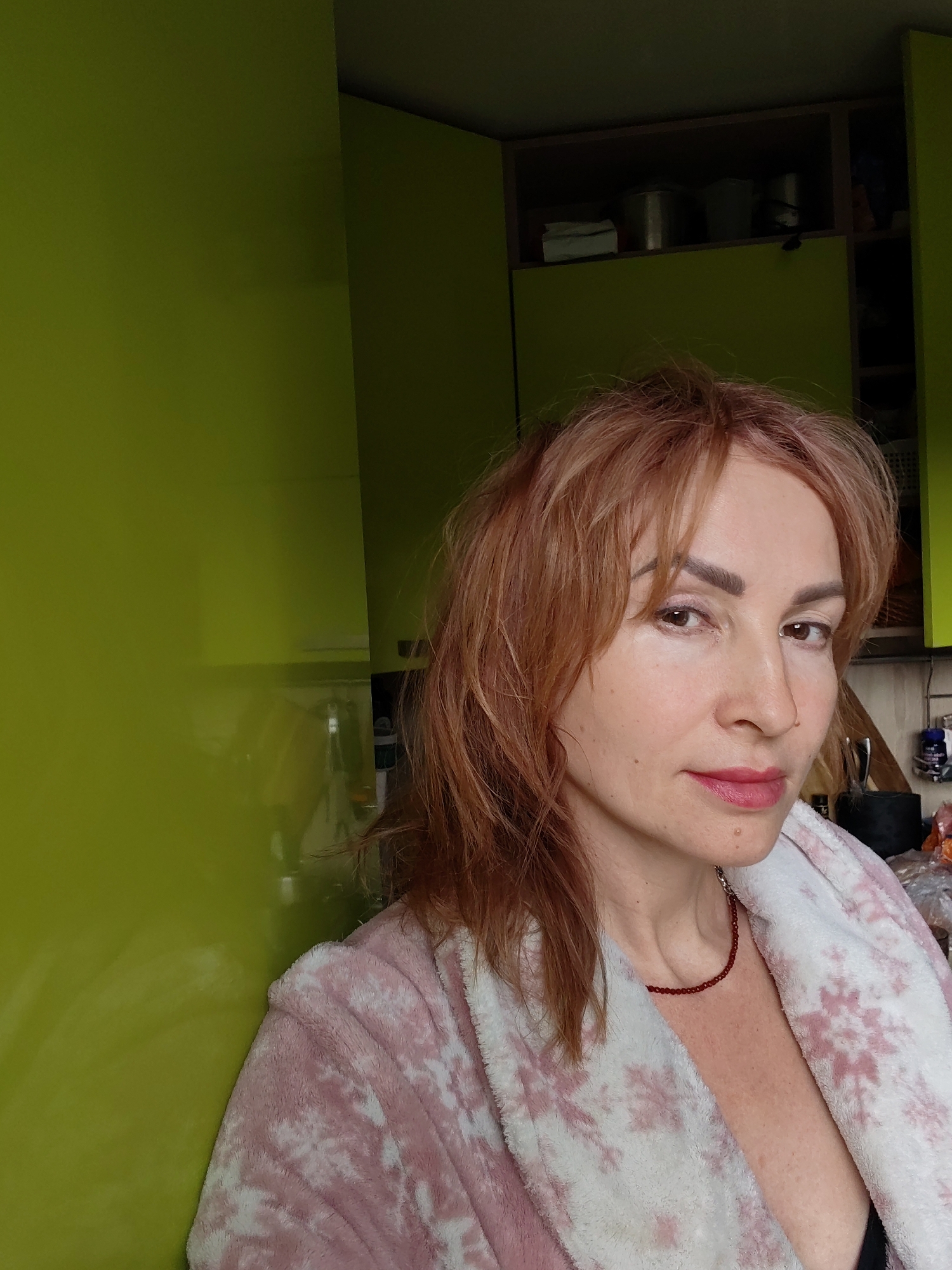 A woman stands indoors, facing the camera at an angle, in what appears to be a kitchen. She has reddish-brown, shoulder-length hair and fair skin. She is wearing a pink and white bathrobe and a red beaded necklace. The background includes bright green kitchen cabinets. There are objects visible in the cabinets. The photograph was taken in Chaikovsky, Russia.