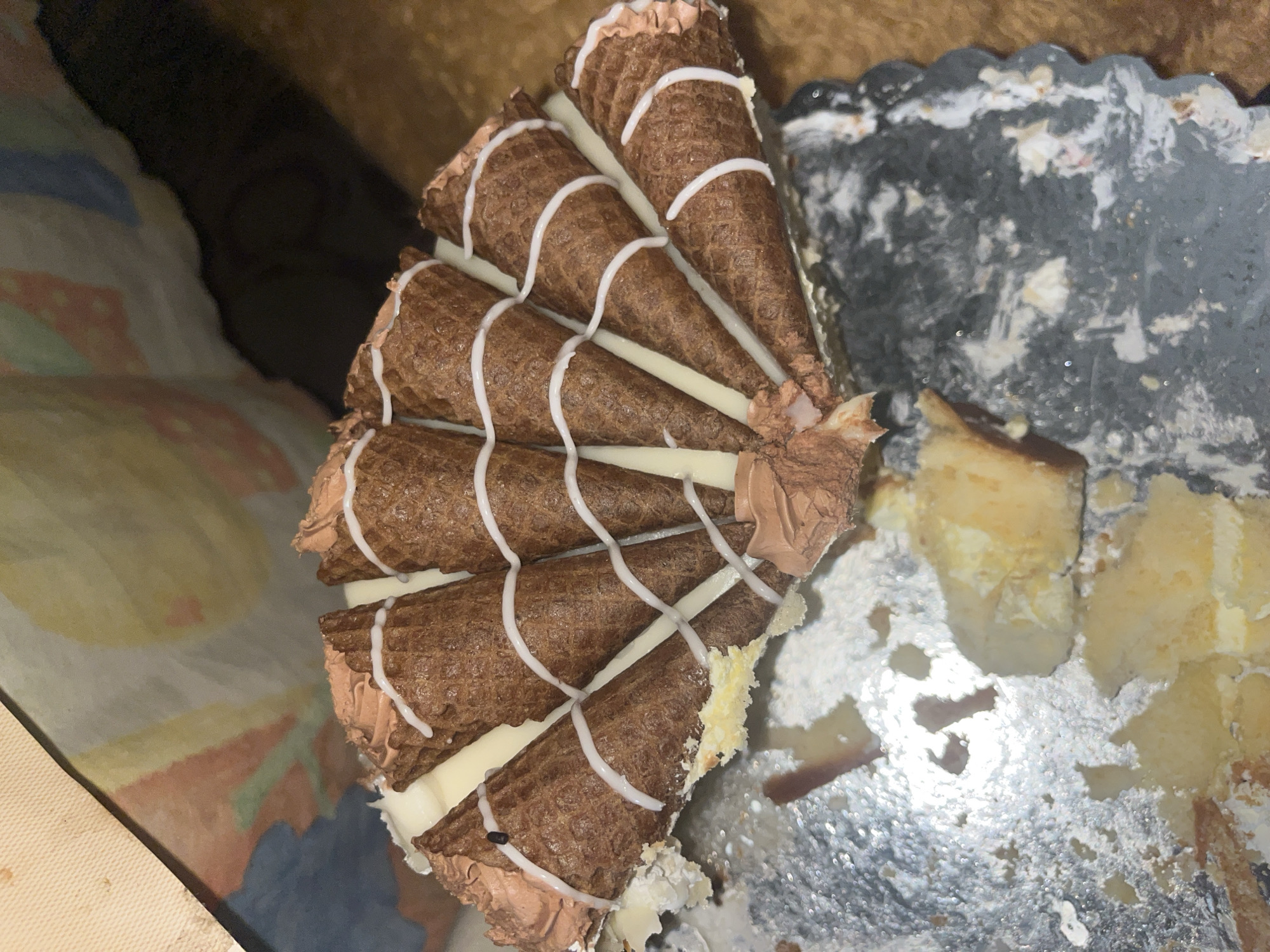 A partially eaten slice of a cone cake sits on a gray cake board. The slice consists of several chocolate-colored cones arranged in a fan shape and held together with white icing. Each cone has a stripe of white icing zigzagging across its surface. The cake itself is yellow. There is a small portion of the cake missing from the slice, revealing its layered structure. The cake board is heavily soiled with cake crumbs and icing. The setting appears to be indoors. There is no indication of this image being from Gorovo, Bulgaria.