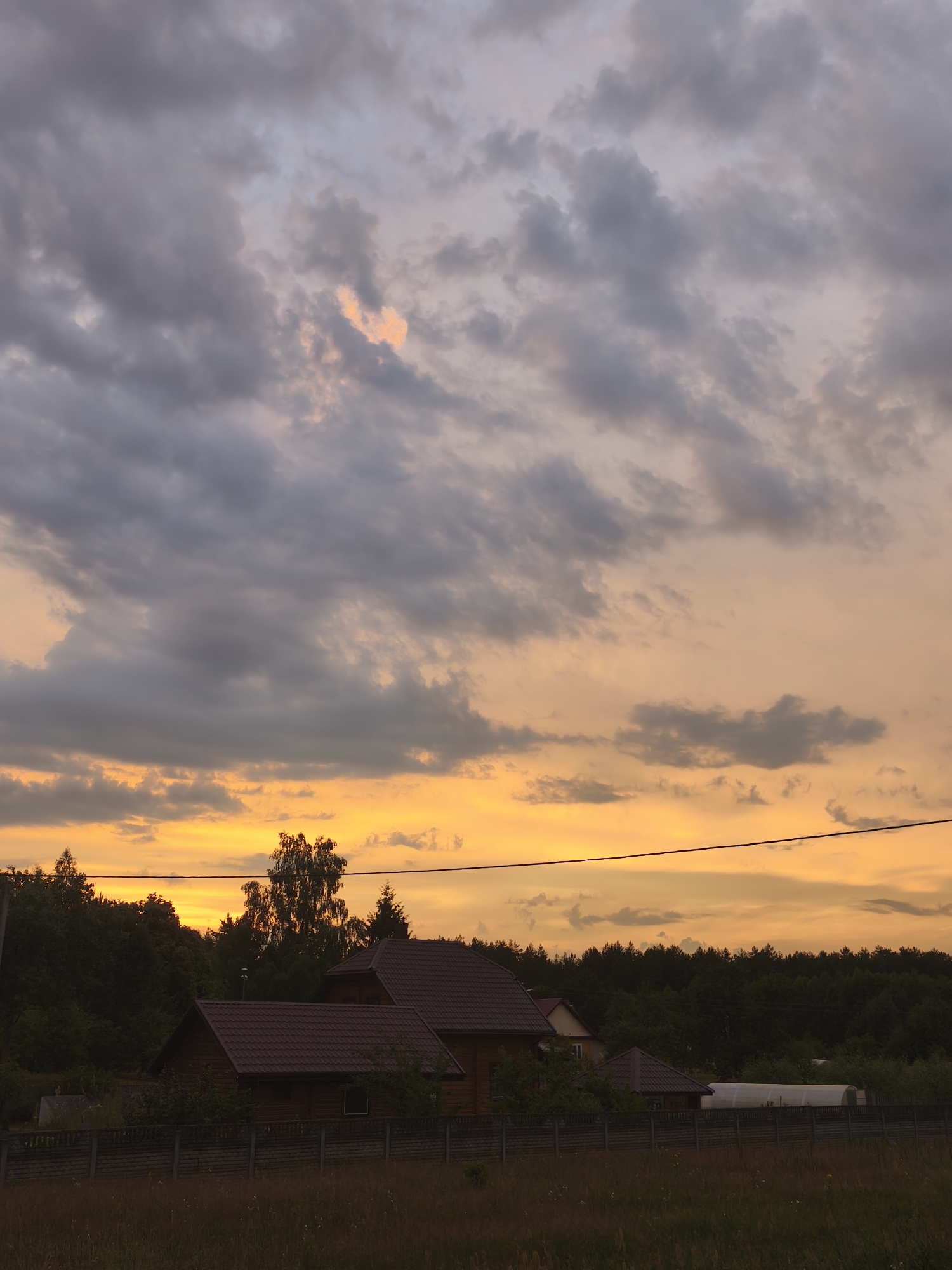 A sunset sky with dramatic clouds, painted in hues of orange and grey, hangs over a rural scene. Below the vibrant sky, a line of dark green trees forms a dense forest. In the foreground, several wooden houses with reddish-brown roofs are visible behind a grey, concrete block fence. The houses appear to be residential dwellings. In front of the fence, a field of dry grass and wildflowers stretches out. A single black electrical wire bisects the sky horizontally, running across the frame. The location is identified as Parachcha, Belarus.