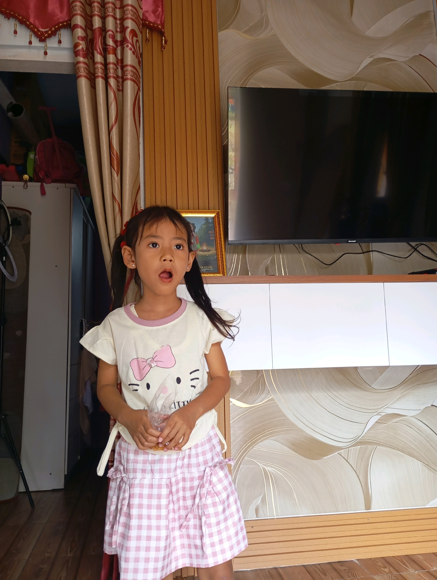A young girl stands in a room, looking surprised with her mouth open. She has dark hair pulled into two pigtails and is wearing a white t-shirt with a pink bow and a cat design, and a pink and white checkered skirt. She holds a clear plastic bag containing some yellow items in her hands. Behind her, a large black television is mounted on the wall above a white cabinet. The wall has a textured wallpaper with wavy patterns in shades of beige and gold. To the left, a doorway is visible, with a red curtain and some items on shelves. The lighting suggests it is daytime. The scene appears to be indoors, possibly in a home in Muncang, Indonesia.