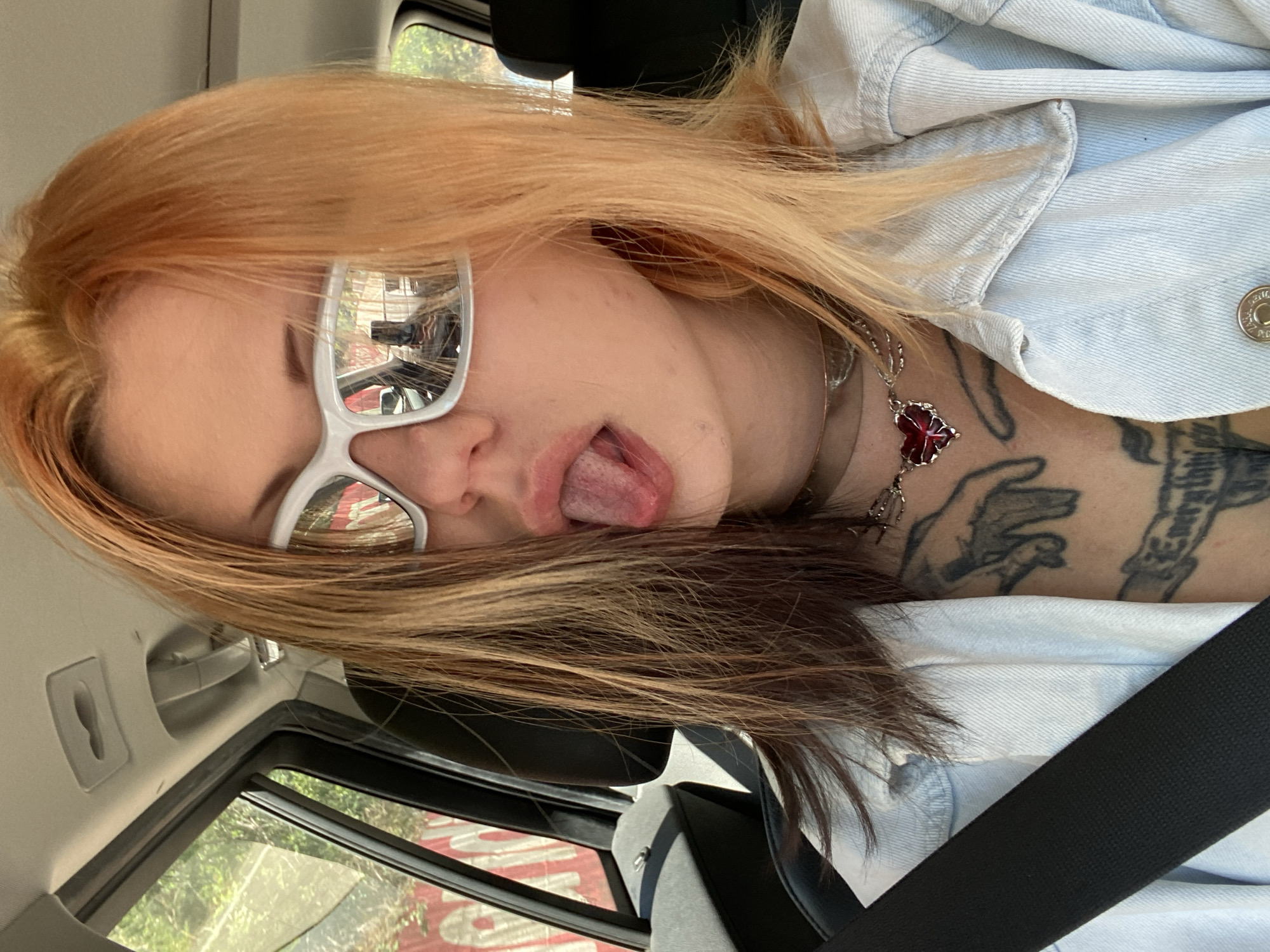 A young person with blonde and brown hair, wearing white-framed glasses and a light blue denim jacket, is shown from a close-up, tilted perspective. The person is sticking their tongue out. The person has tattoos on their neck and collarbone, and a silver necklace with a red pendant. They appear to be in a car, wearing a seatbelt. The reflection in the glasses shows a building and trees outside, possibly in Kruče - Kërruçi, Montenegro.