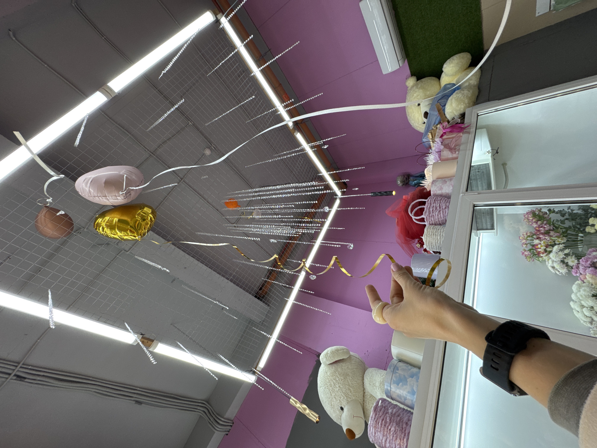 The image shows the interior of a shop or establishment with a high ceiling featuring a gray grid and linear fluorescent lighting. Several helium balloons in bronze, pink, and gold colors are tethered to the ceiling. Decorative elements, including hanging crystal strands and gold ribbons, are suspended from the grid.
The shop's walls are painted in shades of pink and gray. Along one side of the frame, a display of merchandise is visible, including soft plush teddy bears, flowers, and other decorative items.
The foreground contains a portion of a person's arm, adorned with a black sports watch and a ring. The hand appears to be holding one of the gold ribbons.
No specific details in the image suggest the location is Sochi, Russia.