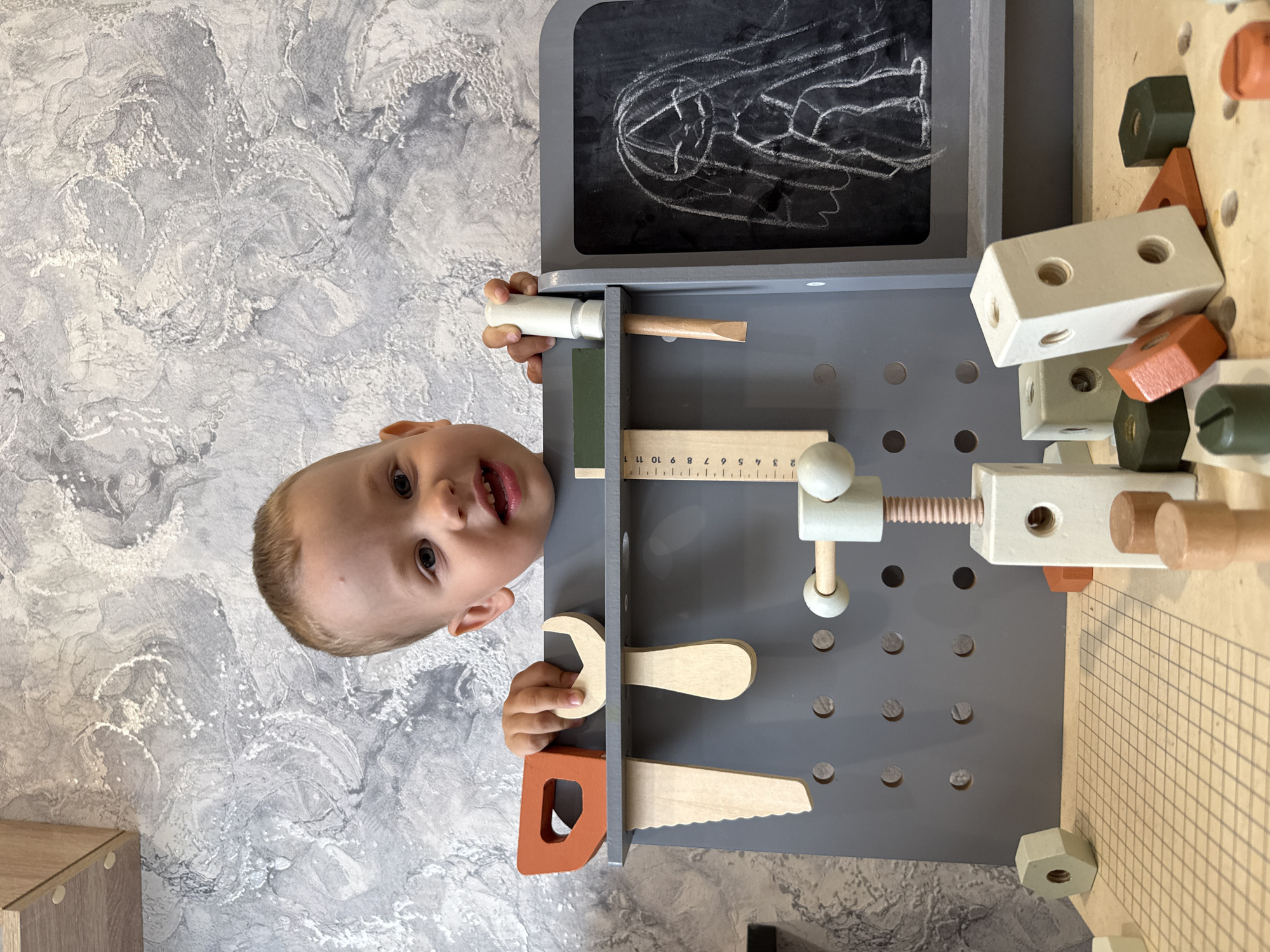 A young child is peering over the top of a grey wooden toy workbench. The workbench has a variety of wooden tools, including a saw, wrench, ruler, hammer, and several blocks. A small chalkboard is attached to the top of the workbench with a drawing on it. The background is a grey and white textured wall. The location is Rusanovo, Russia.