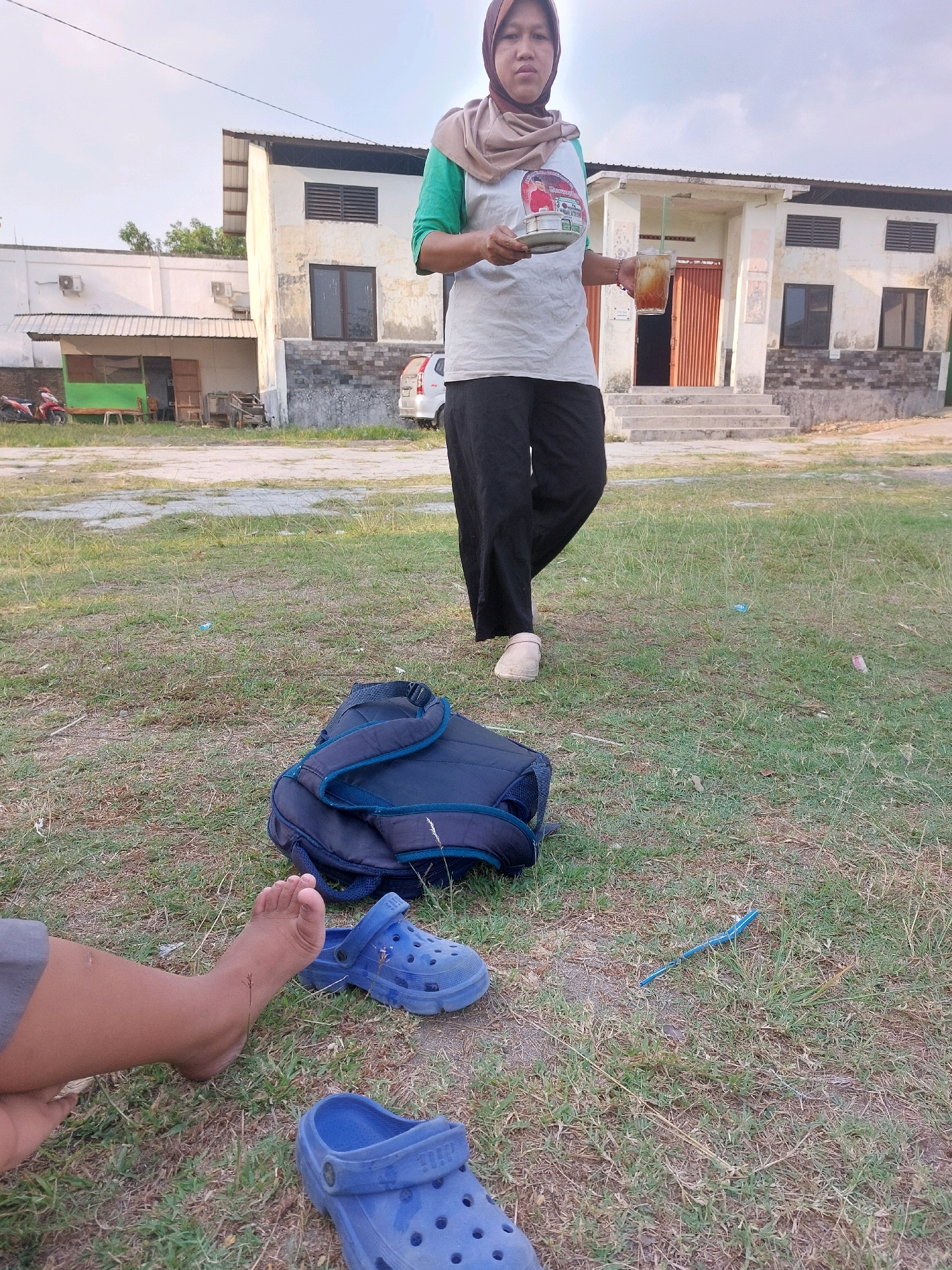 An outdoor scene in SARANGAN, Indonesia features a woman wearing a hijab and a green and white t-shirt, holding a glass of iced tea and a saucer. She is walking away from the viewer. In the foreground, a blue backpack lies on the grass, with a pair of blue Crocs on either side of it. A person's bare foot is visible near the backpack, and another pair of blue Crocs is also present. In the background, a building with a light-colored facade and dark shutters stands under a clear sky. A red motorcycle is parked to the left of the building.