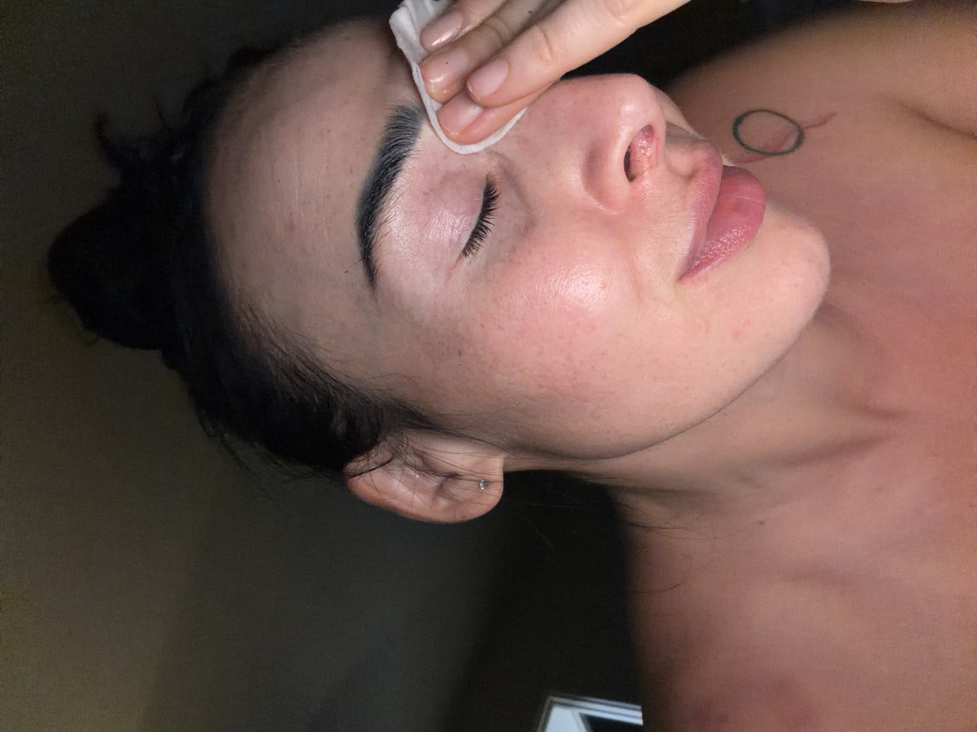 A person is lying down with their eyes closed, receiving skincare treatment. A hand is using a cotton pad to cleanse the area above the person's eyebrow. The person has dark hair pulled back, and visible skin details such as freckles and a small tattoo on their chest. The background is muted and appears to be indoors. The location is not discernible from the image.