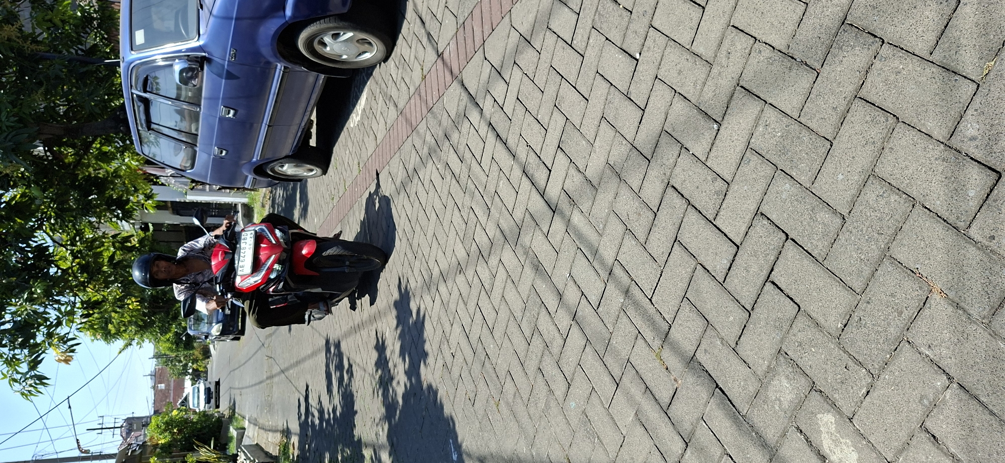 A person wearing a black helmet and a patterned shirt rides a red and black scooter down a street paved with grey interlocking bricks. The scooter has a license plate that reads "AE 6448 JBD". To the right of the scooter, a blue minivan is parked. Green trees and foliage line the street, and overhead, power lines are visible against a clear blue sky. The sun casts shadows of the trees and power lines onto the brick pavement. In the background, houses and other vehicles can be seen. This scene appears to be in a residential area of Surabaya, Indonesia, given the architectural style of the houses and the type of vehicles present.