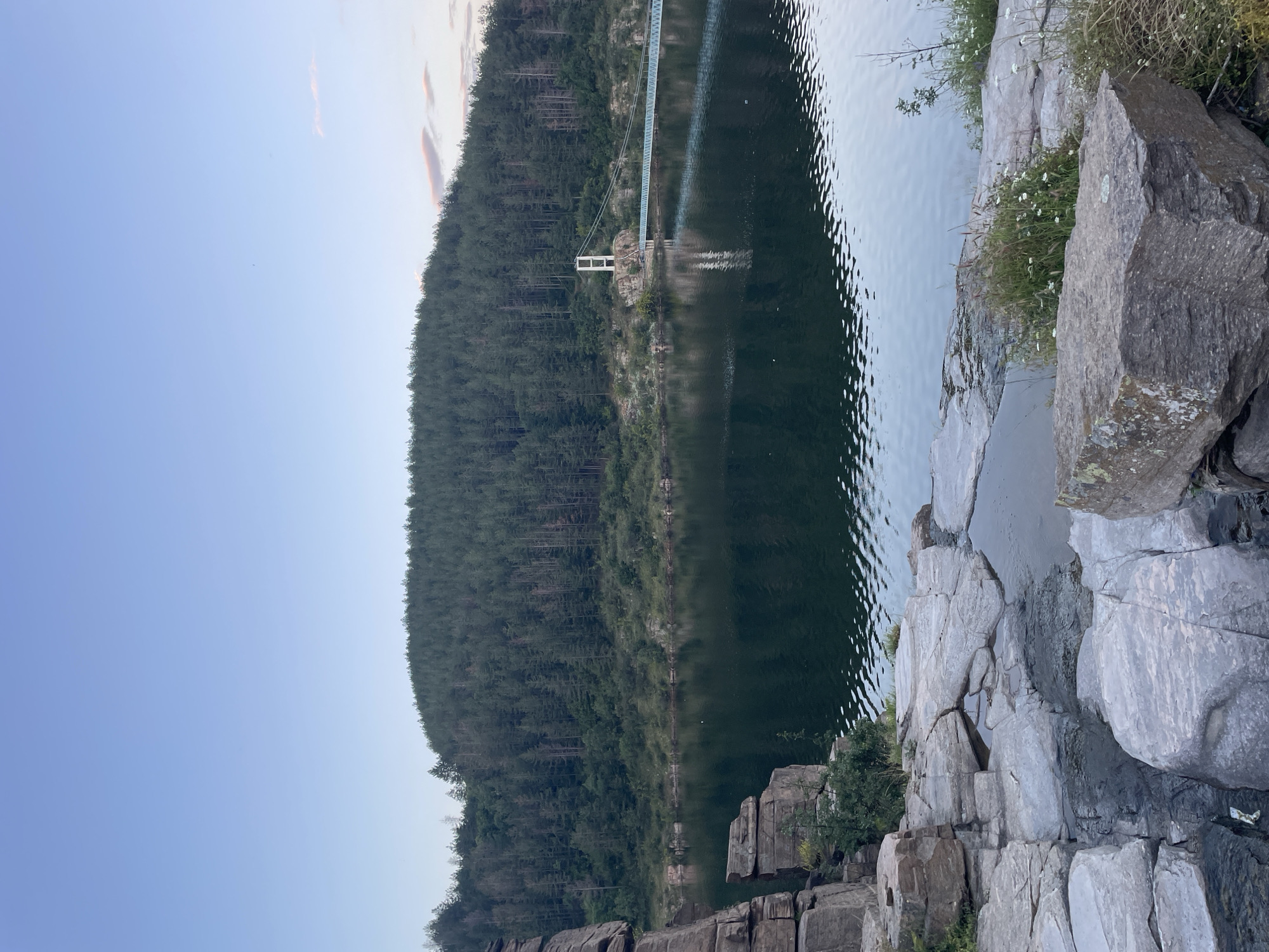 The image depicts a serene natural landscape at dusk. In the foreground, large, weathered rocks form a rugged embankment leading to a calm body of water. The water's surface reflects the surrounding greenery and the sky, with gentle ripples indicating a slight breeze.
Dominating the mid-ground is a dense forest covering a hillside, its dark green foliage stark against the fading light. Across the water, a modern suspension bridge spans the distance, its white support structure and light blue walkway a contrasting element of human engineering within the natural setting. The bridge appears to be a prominent feature of the area.
In the distance, the sky is a soft gradient of pale blue, with hints of pink from a setting sun or approaching twilight. The overall atmosphere is tranquil and picturesque.
This scene is likely located at the Batak Dam, which is situated in the Rhodope Mountains, a region that includes areas near Suhovo, Bulgaria. The specific geographical features, including the rocky shoreline, the forested hills, and the suspension bridge, are characteristic of the reservoir and its surroundings.