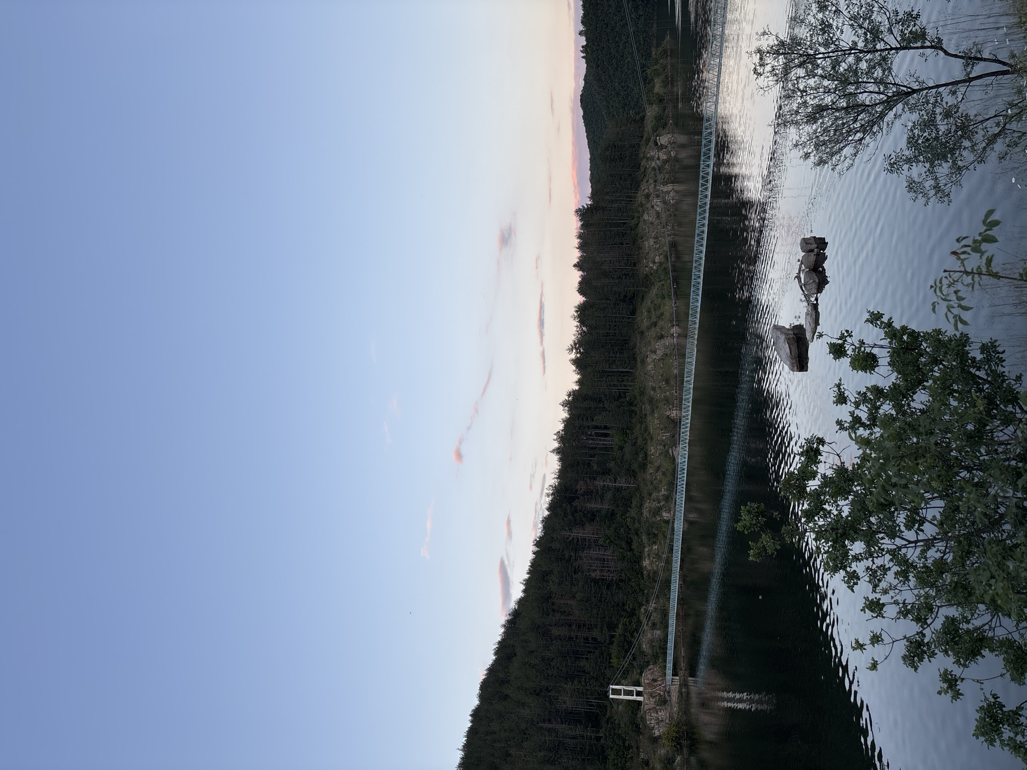 The image depicts a serene lakeside scene at dusk, likely in the vicinity of Suhovo, Bulgaria. A long, slender suspension bridge with a light green railing stretches across the dark, reflective water. The bridge is supported by white pillars on either side, one of which is visible on the left bank. The left bank is covered with a dense, dark green forest that slopes down towards the water, with rocky outcrops visible near the bridge's start. On the right side of the image, a smaller, darker wooded hill rises, its silhouette softened by the fading light. The sky above is a pale blue, transitioning to soft pink and orange hues near the horizon, indicating the time of sunset. A few small, wispy pink clouds dot the sky. In the foreground, the water's surface is disturbed by ripples, with a cluster of rocks partially submerged in the middle ground. Green foliage from trees and bushes frames the bottom of the image. There are no people or animals visible in the scene.