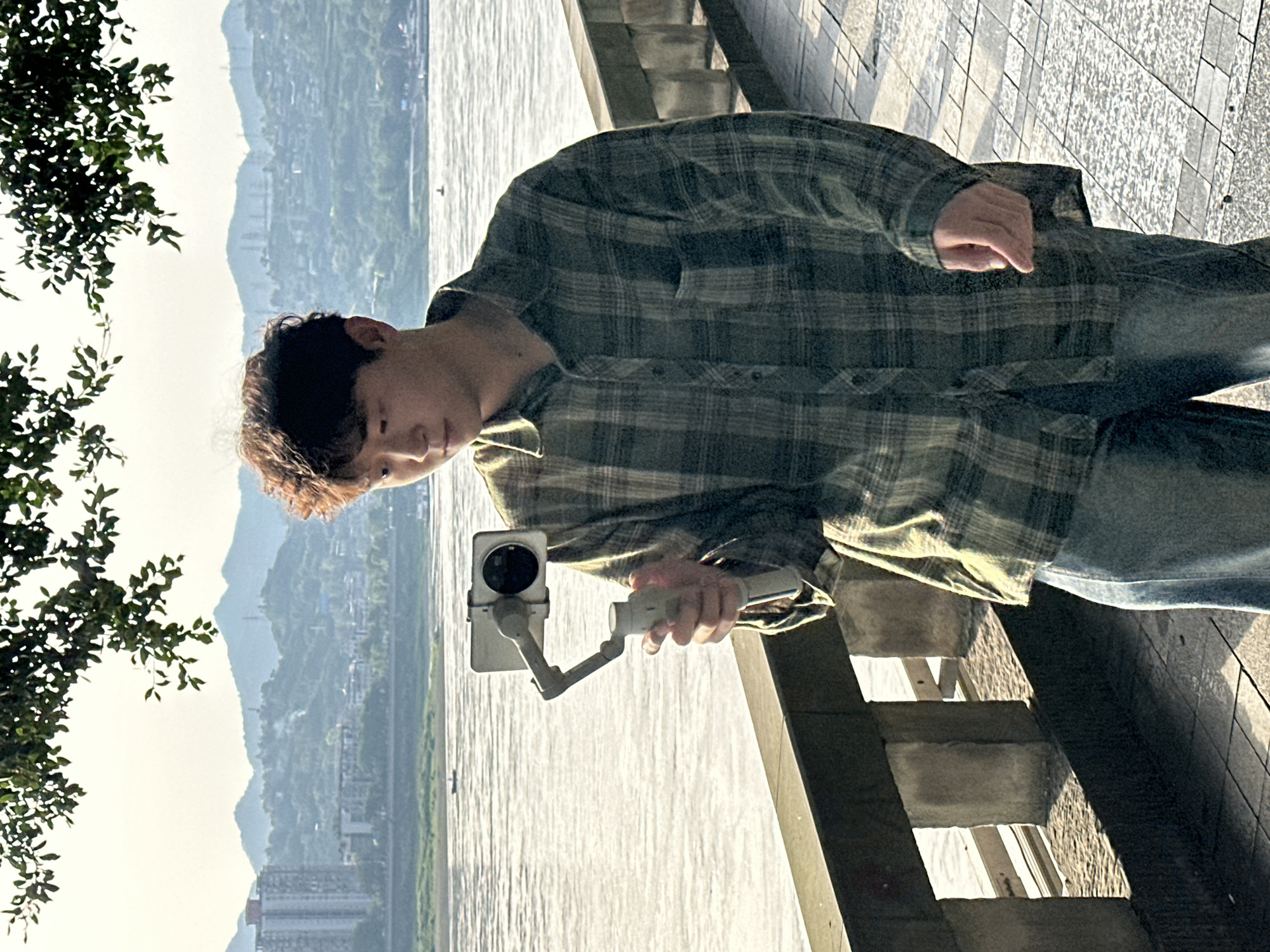 A young man stands on a promenade, likely in Jiangjin, China, holding a smartphone-mounted gimbal for filming or photography. He is positioned near a body of water, possibly the Yangtze River, as suggested by the expansive waterway, distant hills, and city infrastructure in the background. The promenade has concrete stairs and tiling along the edge. The man is wearing a green and grey plaid shirt and blue jeans. He is focused on the phone and gimbal device he is holding. Branches from a nearby tree are visible in the left corner of the frame.
