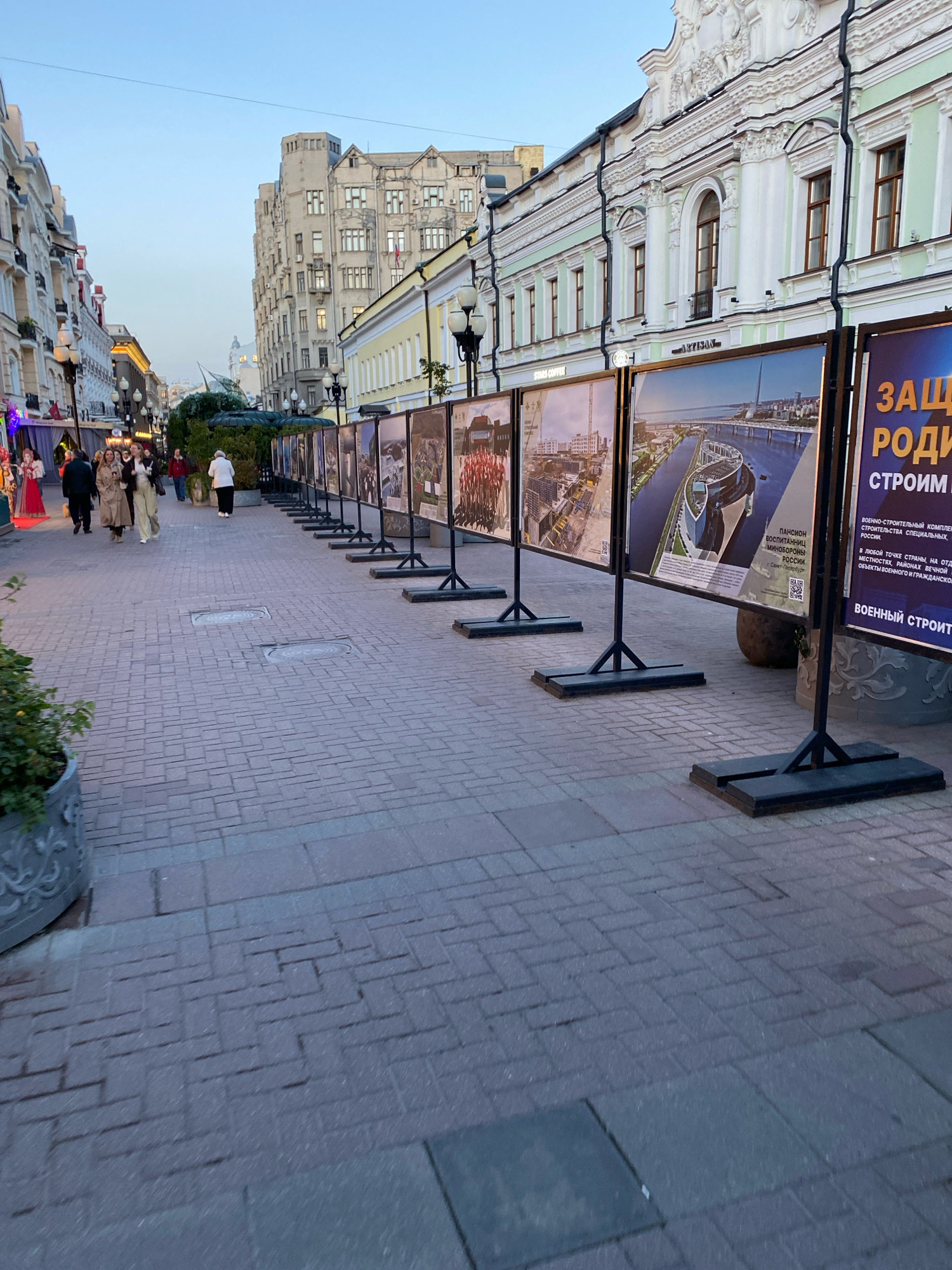 A street exhibition is taking place on a wide pedestrian walkway paved with brickwork, possibly in a historic district of Moscow, Russia. The exhibition features a row of large framed photographs displayed on black metal stands. The photographs appear to depict architectural projects or construction sites, with one image clearly showing a modern stadium structure next to a body of water. Text is visible on some of the photographs, including Cyrillic lettering. In the background, there are multi-story buildings with ornate facades, typical of older European architecture, and streetlights. Several people are walking along the street, some are looking at the photographs. The sky is clear and a pale blue, suggesting it is either early morning or late evening.