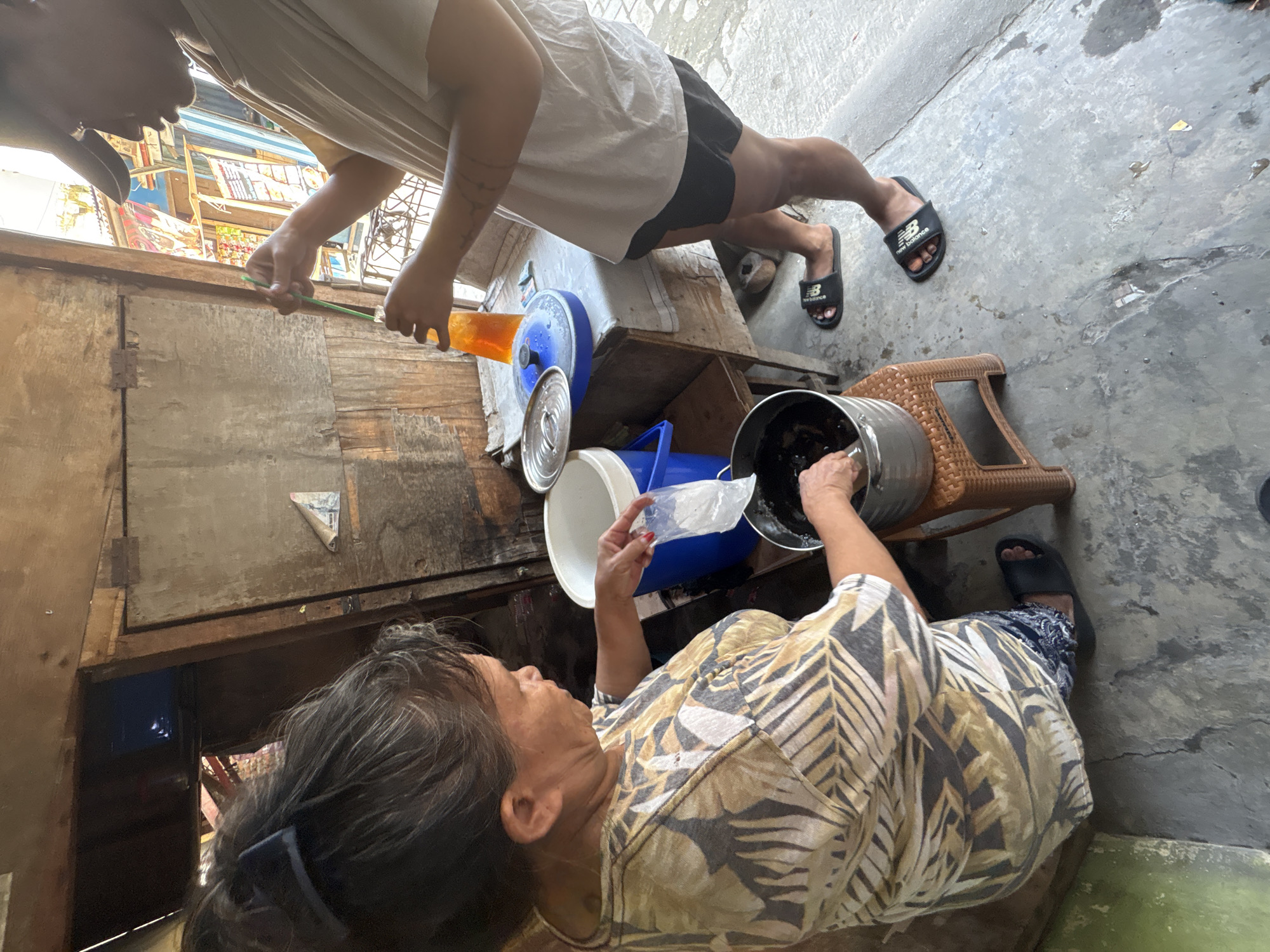In North Jakarta, Indonesia, a scene unfolds at what appears to be a roadside stall. A woman, her hair pulled back with a dark clip, wears a patterned shirt and is focused on a stainless steel pot. In her left hand, she holds a small plastic bag, seemingly pouring its contents into the pot. Nearby, a blue cooler is visible. To the right, a younger person wearing a baseball cap, a light-colored t-shirt, black shorts, and black sandals, is engaged with a tall plastic bottle filled with an orange liquid, stirring it with a green straw. The background reveals a rustic wooden structure and the urban environment of a street. The ground is concrete, showing signs of wear and some dampness. A small dog can be seen peeking from behind a wooden stool.