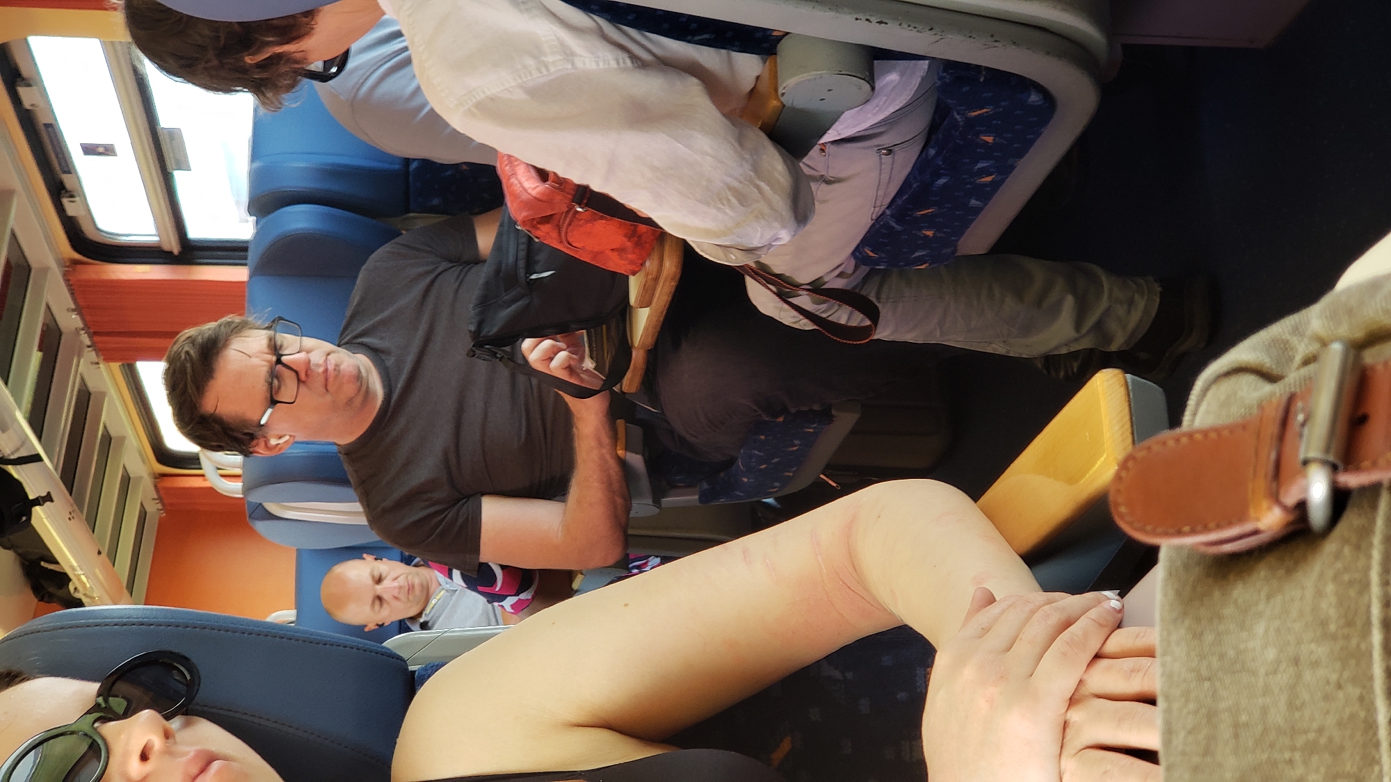 The image depicts an interior view of a train carriage with passengers. In the foreground, the left arm and hand of a person wearing a black top and sunglasses are visible. To the right of this person, a man with glasses and earbuds is seated, looking down. Further back, another man with a bald head and earbuds is also seated. A red bag and a black bag are placed on a tray table in front of the man with glasses. The seats are blue with an orange and blue pattern. The train carriage has orange curtains and windows. The flooring is dark blue. This scene appears to be from a train traveling through or near Horná Streda, Slovakia, given the context.