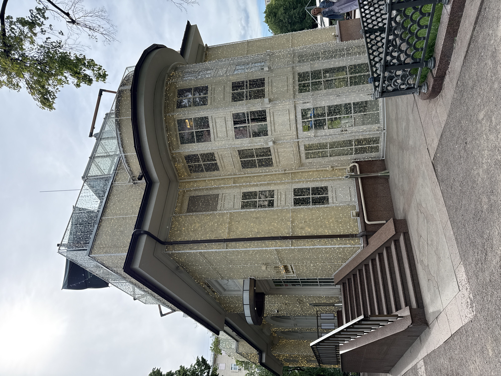 The image displays a multi-story building extensively decorated with numerous small, warm-toned string lights covering its entire facade and a glass-covered upper level. The building features a light-colored, possibly cream or off-white, exterior with rows of rectangular windows and a distinctive curved architectural section.
In the foreground, a wide, dark-toned stone staircase with a robust, decorative black metal railing ascends towards the building's entrance. The ground around the staircase is paved with a mix of light stone tiles and darker concrete. On the left side of the frame, green foliage from a tree is visible against an overcast sky. To the right, beyond the staircase and pavement, there's a decorative metal fence with a scalloped pattern, separating the walkway from a small patch of grass. A person wearing a light-colored jacket or top is partially visible behind this fence in the upper right portion of the image.
The weather appears to be overcast, with no direct sunlight, yet the decorative lights are illuminated. The overall impression is that of a well-maintained urban setting, possibly showcasing seasonal or permanent festive decorations. No visible text can be identified in the image. The location context provided is Moscow, Russia.