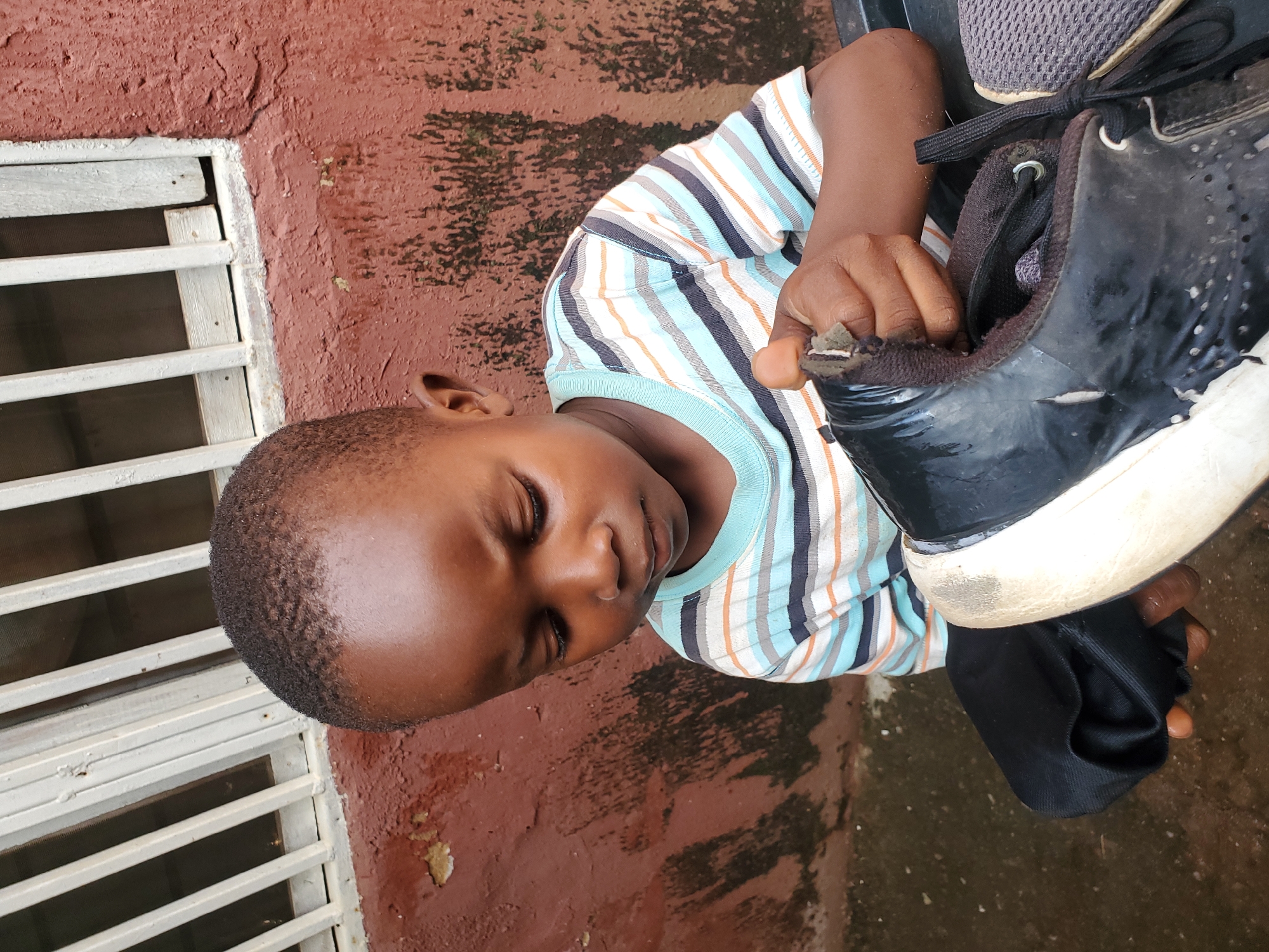 The media file shows a close-up, rotated view of a young Black child sitting on the ground. The child is wearing a horizontally striped t-shirt with colors of blue, white, black, and orange. The child's eyes are closed, suggesting they may be sleeping or resting. One of the child's hands is holding a black sneaker, with their fingers partially inside the shoe. Another shoe, a gray, mesh-like sneaker, is visible in the upper right portion of the image, partially obscured.
The child is positioned next to a rough, reddish-brown wall, which appears textured and somewhat weathered, with some dark green or black splotches, possibly moss or dirt. To the left of the child and the wall, there is a white metal grate or window grill, with horizontal bars. The ground appears to be wet or damp, with some small, light-colored debris or stains visible on the reddish surface.
The overall scene suggests an outdoor setting, possibly in a residential area or public space. The lighting does not strongly indicate a specific time of day. The child's closed eyes and relaxed posture do not convey any strong emotions. There is no visible text in the image. The context of Jos, Nigeria, suggests a potentially warm climate, which might explain the child's attire and outdoor setting.