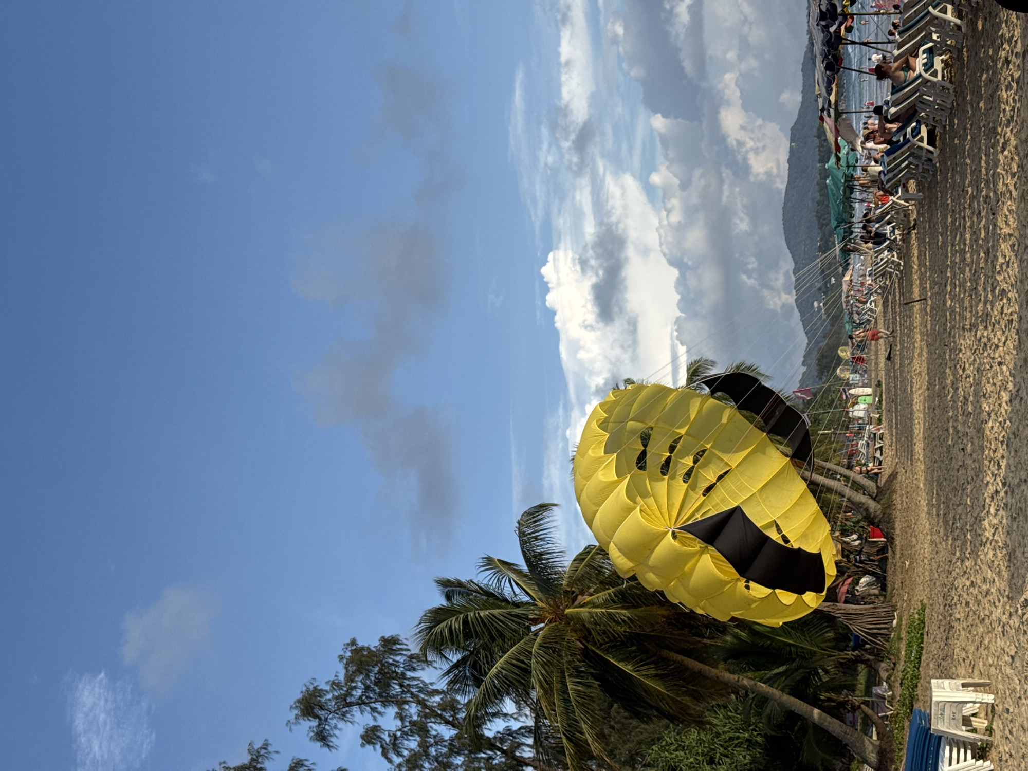 This image depicts Karon Beach in Karon, Thailand, under a bright blue sky with scattered clouds. In the foreground, a large yellow and black parachute for parasailing dominates the left side of the frame, partially obscuring a cluster of palm trees. The beach itself is sandy, with rows of white lounge chairs and green umbrellas set up for sunbathers. Numerous people are visible relaxing on the chairs, walking along the shore, and swimming in the ocean. The background features a lush, tree-covered mountain range. The overall scene suggests a lively and popular tourist destination.