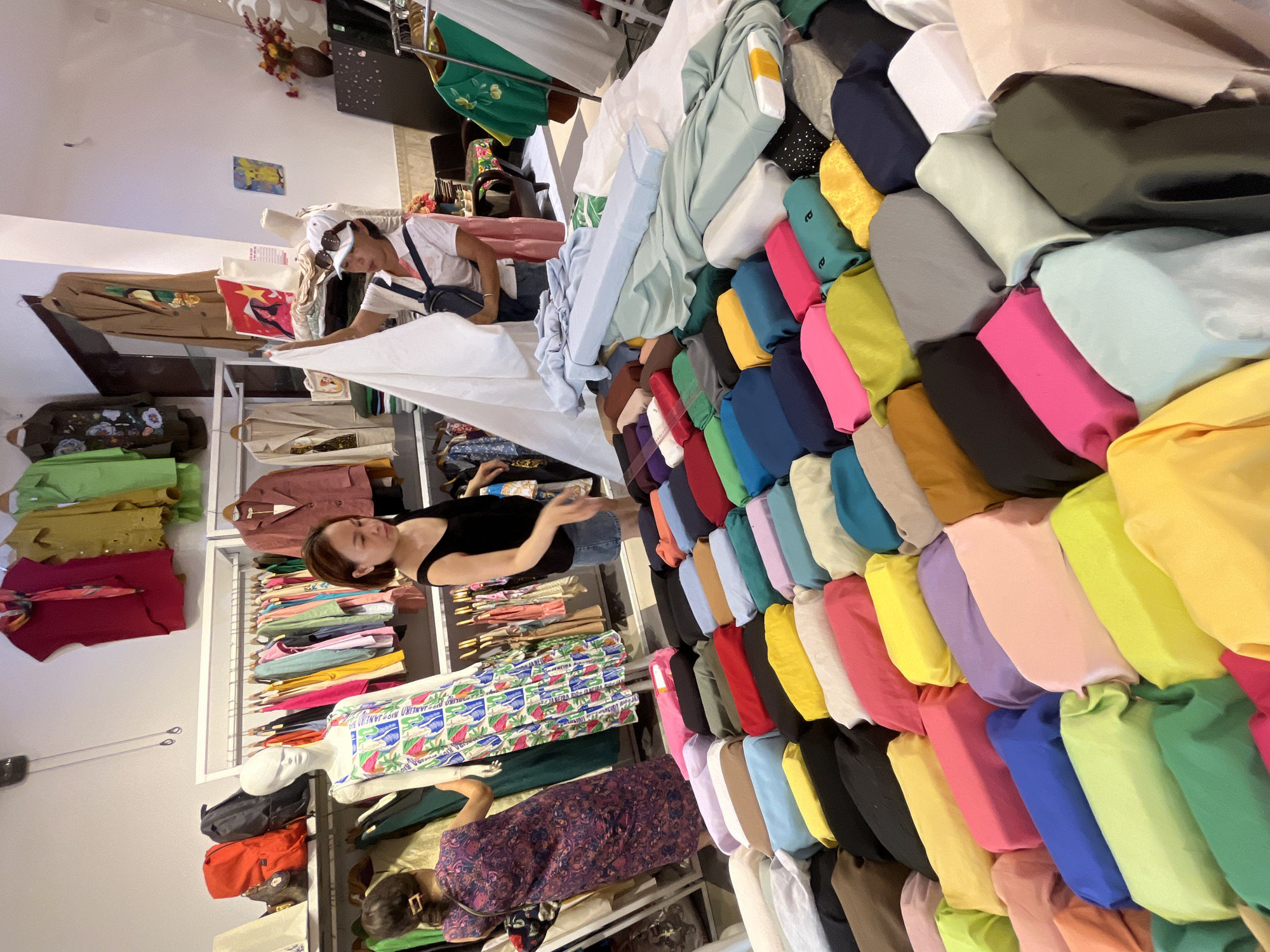 A busy clothing store in Da Nang, Vietnam, is filled with racks of colorful garments, bolts of fabric, and mannequins. Two women are seen interacting with the merchandise. One woman, wearing a purple patterned dress, is standing and looking at clothing on a rack. Another woman, in a black tank top and denim shorts, is holding a white piece of fabric, seemingly examining it. A third woman, wearing a white shirt, sunglasses, and a baseball cap, is also holding fabric. The store appears to be well-stocked with a variety of clothing and textiles, suggesting it is a retail establishment catering to fashion and fabric needs.