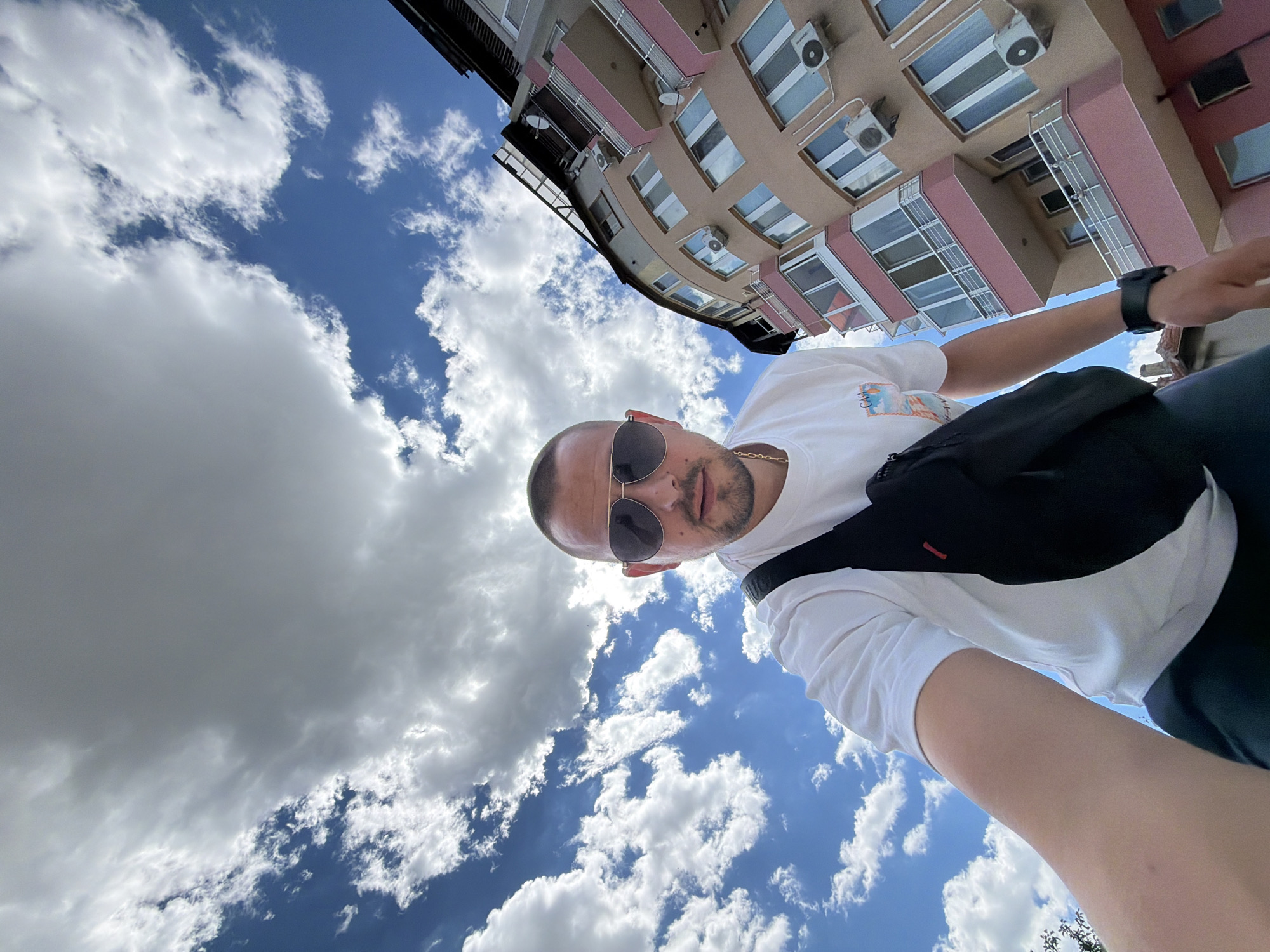 The image is a low-angle, wide shot selfie taken outdoors, with a person in the foreground and a building and sky in the background. The person, a male, is wearing a white t-shirt, sunglasses, a gold chain, and a black fanny pack worn across the chest. He is looking towards the camera. To the right of the person is a multi-story building with a curved facade. The building is painted in shades of beige and pink, with balconies, windows, and air conditioning units visible. The sky above is a bright blue with scattered white and grey clouds. A portion of a dark, possibly residential, building is visible on the far right, with a slanted roof. The scene appears to be a street in Burgas, Bulgaria, given the architectural style of the building.