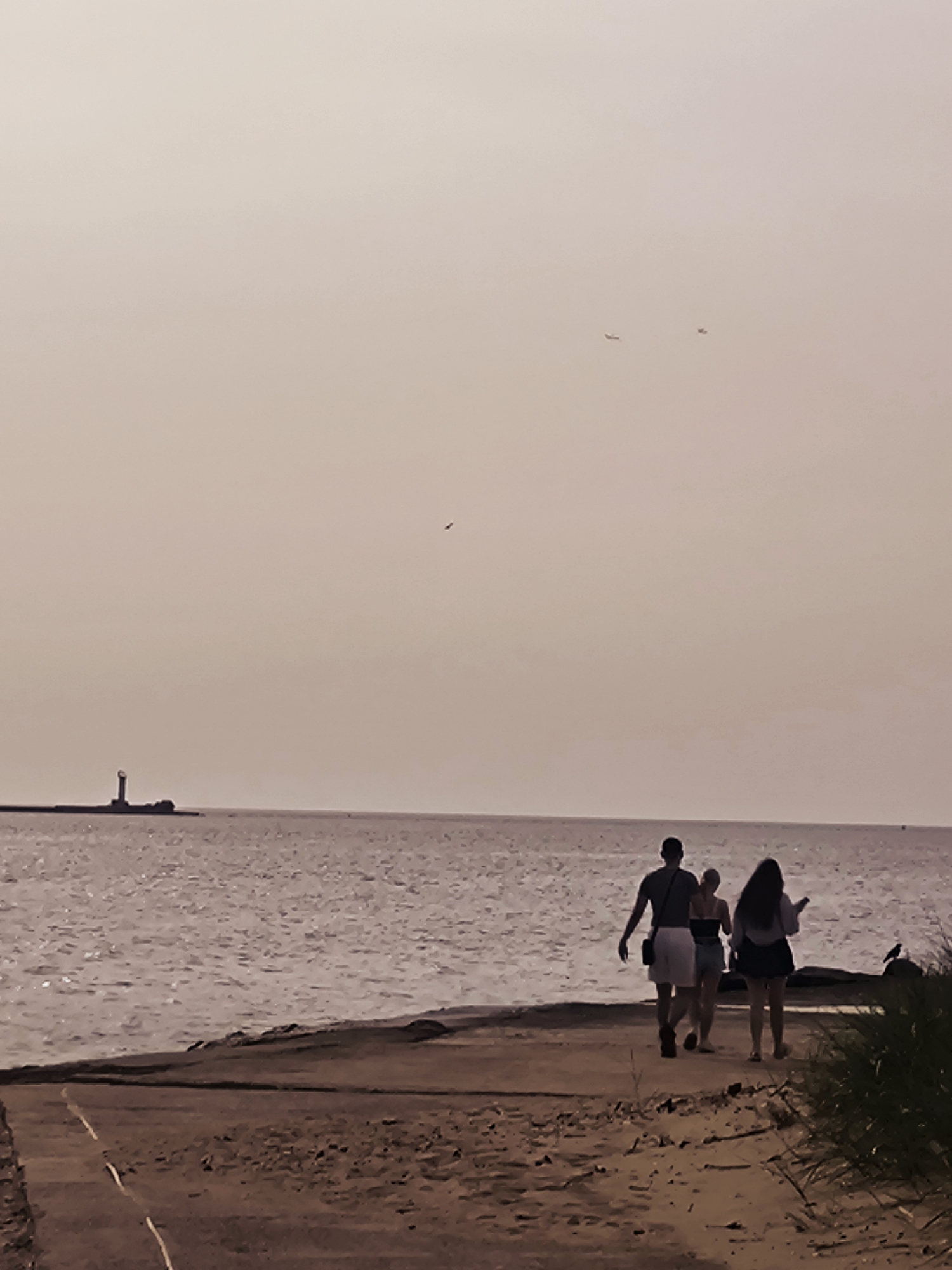 A group of three people are walking away from the camera along a sandy path next to a body of water. The person in front is a male wearing shorts and a t-shirt. Behind him are two females, one in a tank top and shorts and the other in a long-sleeved shirt and skirt. In the distance, a lighthouse stands on a pier extending into the water. The sky is overcast and a muted pinkish-gray. Two small aircraft are visible in the sky. The scene appears to be a beach or coastline. The location is not identifiable as Riga, Latvia from the image.