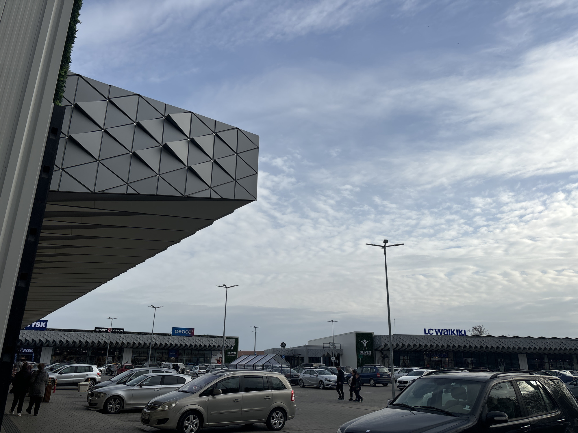 The image shows a parking lot in front of a shopping center in an unknown city. The sky is partially cloudy. The facade of the building is decorated with gray triangular panels. Various cars are parked in the lot. Store signs visible include "JYSK", "SPORT VISION", "pepco", "SLIVEN RETAIL PARK", and "LC WAIKIKI". Several pedestrians are walking in the parking area. Streetlights are interspersed throughout the parking lot.