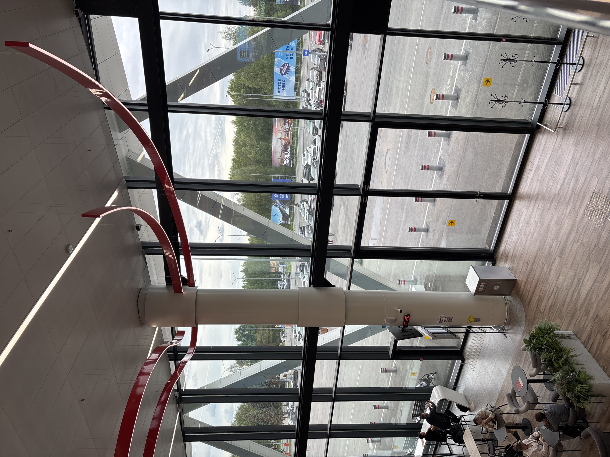 This image depicts an interior view of a modern building with large windows overlooking an outdoor parking lot and a street. The interior features a white cylindrical pillar with red curved decorative elements extending from the ceiling. The floor is made of light brown wooden planks.
Several people are visible inside the building, seated at tables in what appears to be a waiting area or lounge. They are engaged in various activities, such as looking at their phones or talking to each other, suggesting a relaxed or waiting atmosphere.
Outside, the parking lot is filled with numerous cars. Large billboards are visible in the distance, some displaying text. The weather appears to be overcast, with a cloudy sky. The overall scene suggests a public space, possibly an airport or a large commercial building, given the scale of the windows and the presence of parking and billboards. The digital clock on the pillar indicates the time is 17:26. Some text on the billboards is partially visible, including "ЧИСТОЕ НЕБО" (CLEAN SKY) and "ЭРОДЫ КОЛОКОЛА" (EROS OF THE BELL). The location context is Старое Мартьяново, Russia.