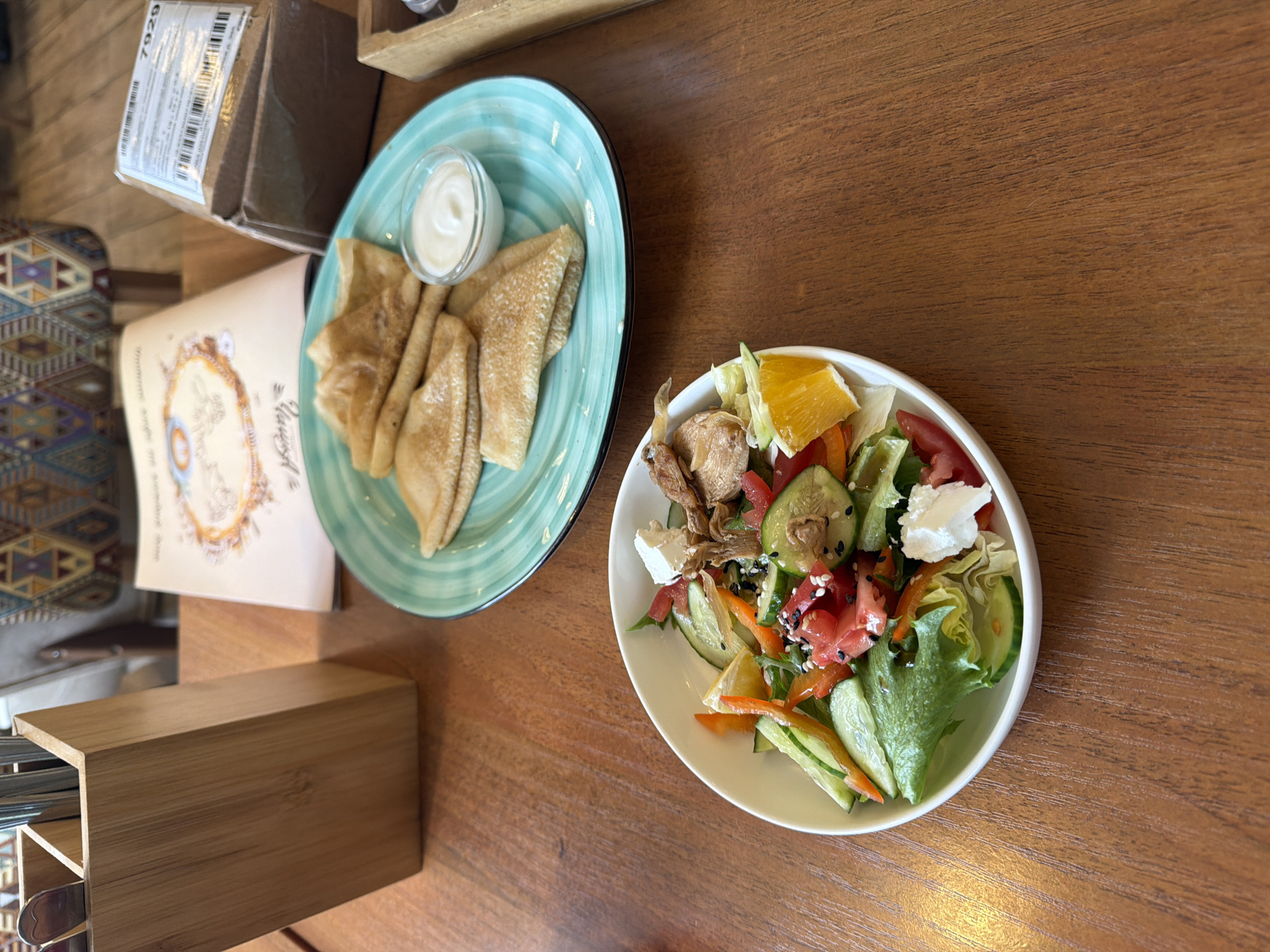 A high-angle medium shot captures a dining scene on a wooden table in Izhevsk, Russia. In the foreground, a white bowl contains a salad with lettuce, cucumber, tomatoes, orange bell pepper, feta cheese, and black sesame seeds. To the right and slightly behind the salad bowl, a turquoise plate holds several folded crepes with a small dish of sour cream. Behind the crepes, a cardboard box with shipping labels and a menu are visible. To the left of the frame, a wooden cutlery holder with utensils stands upright. The background shows a patterned chair and a wall. The lighting is soft and natural, casting subtle shadows. No people are visible in the frame.