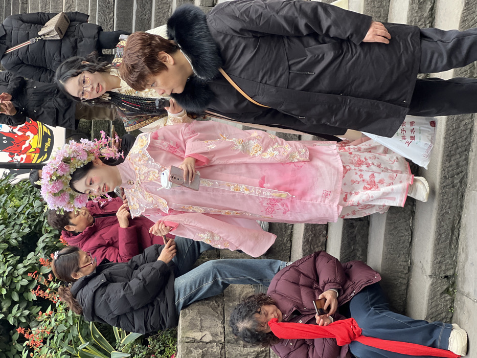The image captures an outdoor urban scene in Yuzhong District, China, primarily focused on a group of individuals on and beside stone steps. In the center, a young woman with dark hair wears a large, elaborate headpiece composed of pink flowers and green foliage. She is dressed in a light pink traditional-style hanfu with intricate gold and pink embroidery on the collar, sleeves, and tunic, over a white floral-patterned skirt. She holds a white disposable cup in her left hand and a light-colored smartphone in her right, facing right with a slight smile.
To her immediate right, an older woman in a black puffer jacket with a prominent black fur-lined collar stands, looking right, out of frame. A white plastic shopping bag with Chinese characters "胖子妈 王婆鸭" (Fatty Mom Wang Po Duck) is visible near her feet. Slightly behind the central figure, a young woman with dark, wavy hair and glasses wears a patterned vest over a light-colored top and holds a small black device, possibly a camera or recorder, pointed towards the central figure.
To the left, on a lower stone step, a woman with dark, curly hair wears a dark red puffer jacket and blue jeans, looking down at a smartphone in her hands while holding a red fabric drape. Behind her, seated on a low stone wall, another woman in a black puffer jacket and blue jeans is also engrossed in a smartphone. Further back, an older woman in a dark red jacket is partially visible. The background on the left features green foliage and some red flowering plants along the stone wall. In the upper background, additional figures are present on the steps, and a partially visible sign with red and black graphics and the word "WHO IS" is mounted on a building. The entire scene is bathed in natural daylight.