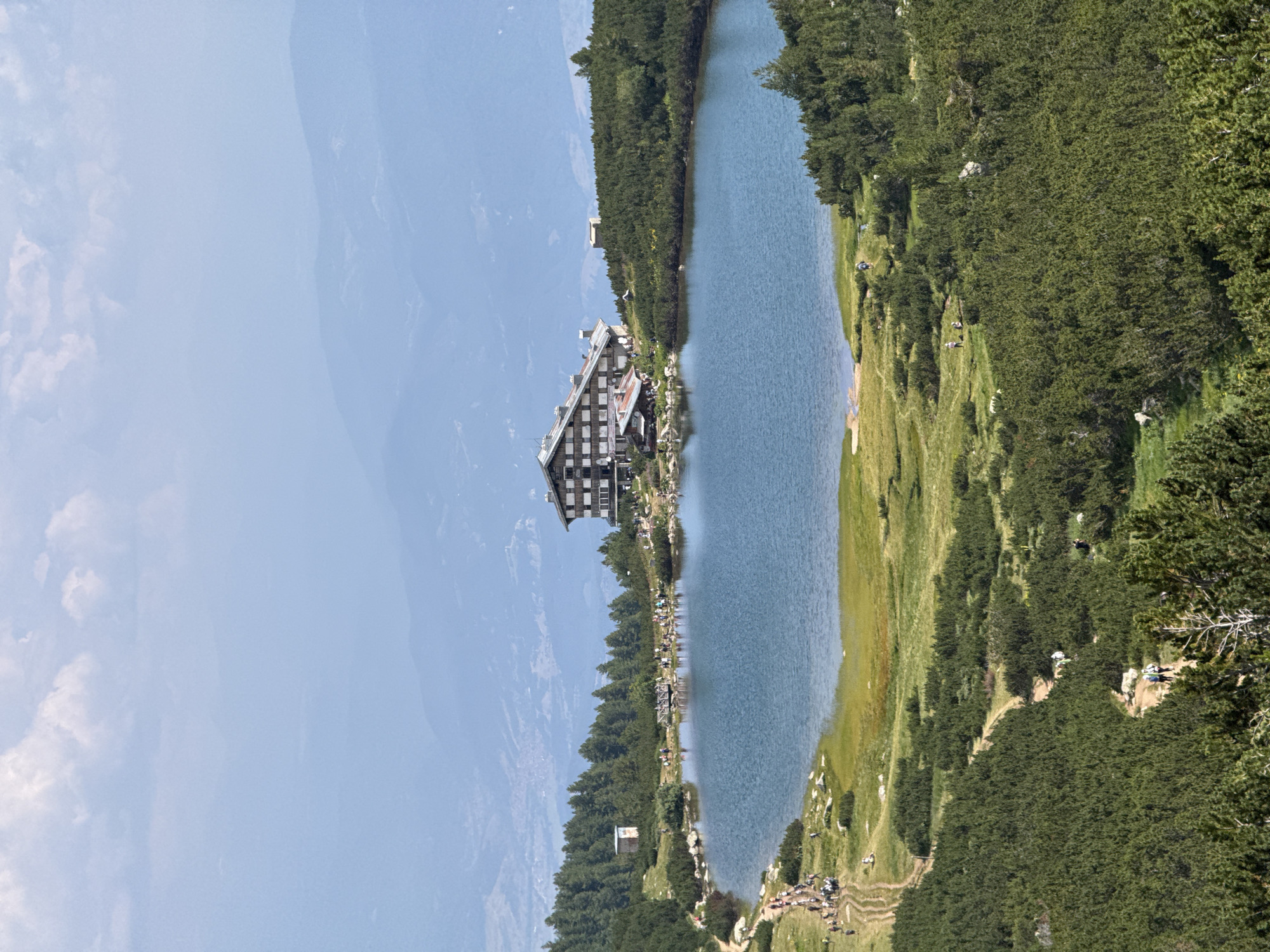 The image depicts a scenic mountain landscape featuring a tranquil lake surrounded by lush greenery and coniferous trees. In the background, a large, multi-story building, possibly a lodge or hotel, is situated on a hillside overlooking the water. Several people are visible around the lake, some walking along the shore or on trails, while others are gathered near the building. The terrain is characterized by rolling hills and distant mountains shrouded in a hazy atmosphere. The foreground is dominated by dense evergreen bushes and trees, with a winding path leading down towards the lake. The sky is partly cloudy, with patches of blue visible. The overall scene suggests a popular tourist destination, likely in a mountainous region, offering opportunities for hiking and enjoying nature. This specific location is identified as the Seven Rila Lakes, located in the Rila Mountains, near the town of Bansko, Bulgaria, a region known for its natural beauty and hiking trails.