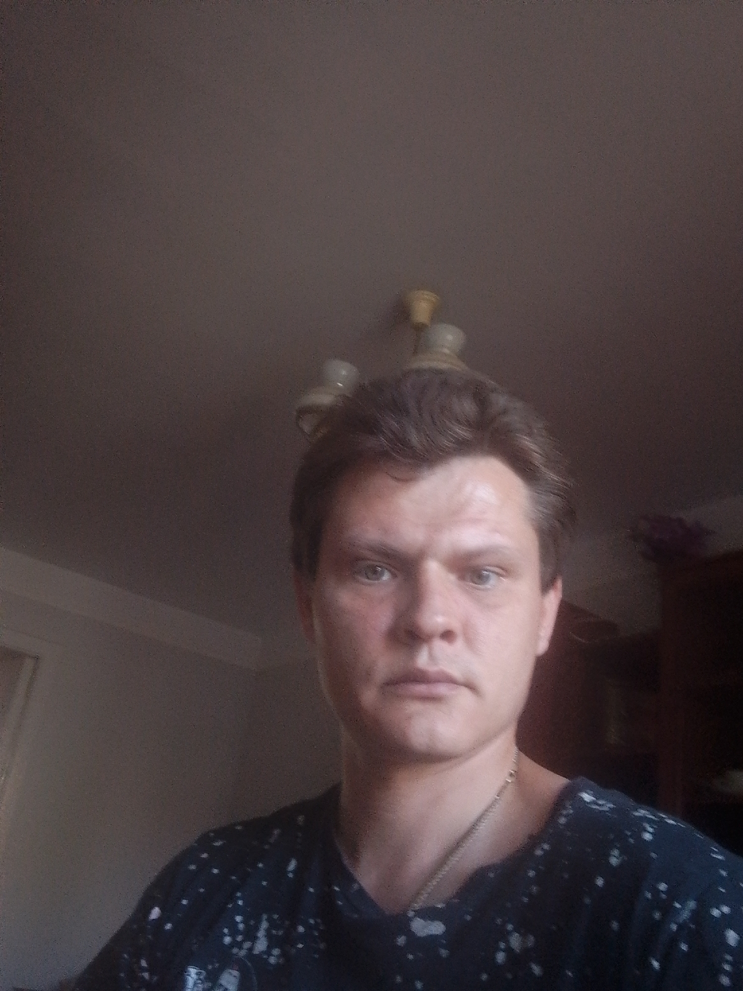 A man is captured in a close-up, waist-up selfie taken from a slightly low angle. He has fair skin, light brown hair, and light blue eyes. He is looking directly at the camera. The man is wearing a dark t-shirt with white speckles and a thin silver chain necklace. The background appears to be an indoor setting, possibly a room, with a plain, lightly colored ceiling and a darker wall behind him. A light fixture is visible on the ceiling. The lighting suggests the light source is above and to the front of the subject.