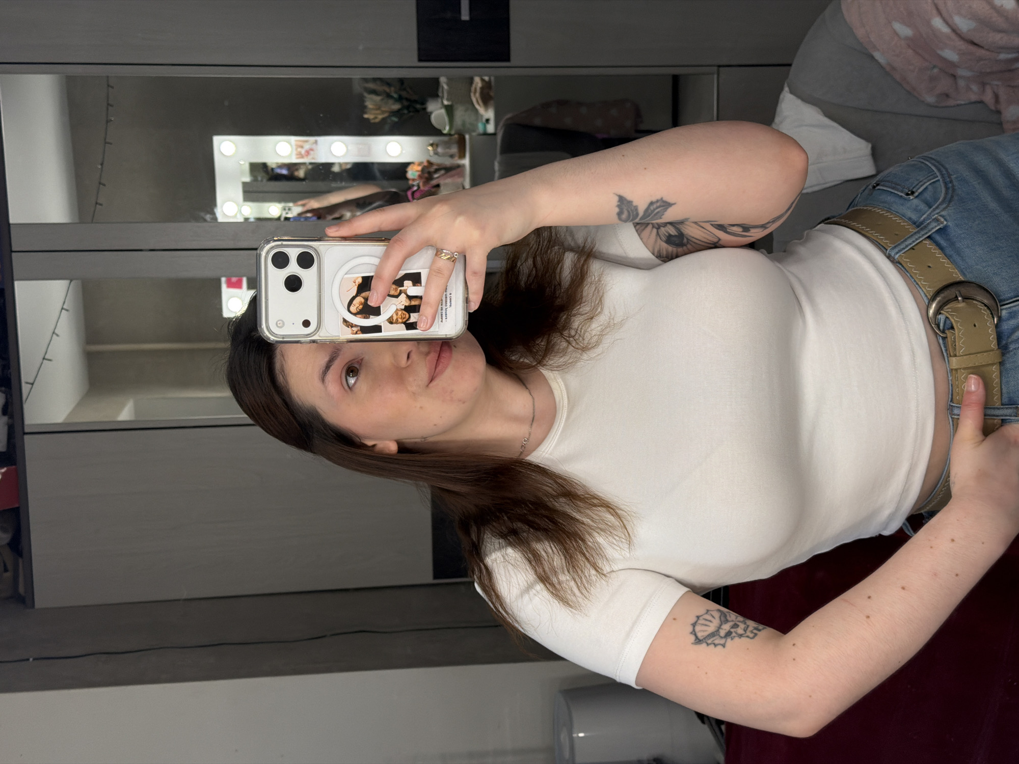 A young woman takes a selfie in front of a mirror in what appears to be an indoor setting. She is wearing a white t-shirt, light blue jeans with a tan belt, and a silver necklace. Her hair is brown and shoulder-length. She is holding a white iPhone with a transparent case featuring an image of the TV series "Friends" on the back. The mirror reflects a vanity with lights and various objects on top. The room appears to be in a state of partial organization. A bed or seating area with a gray and pink patterned blanket can be seen in the background. The location of the city of Chaikovsky, Russia cannot be confirmed with certainty from the image.
