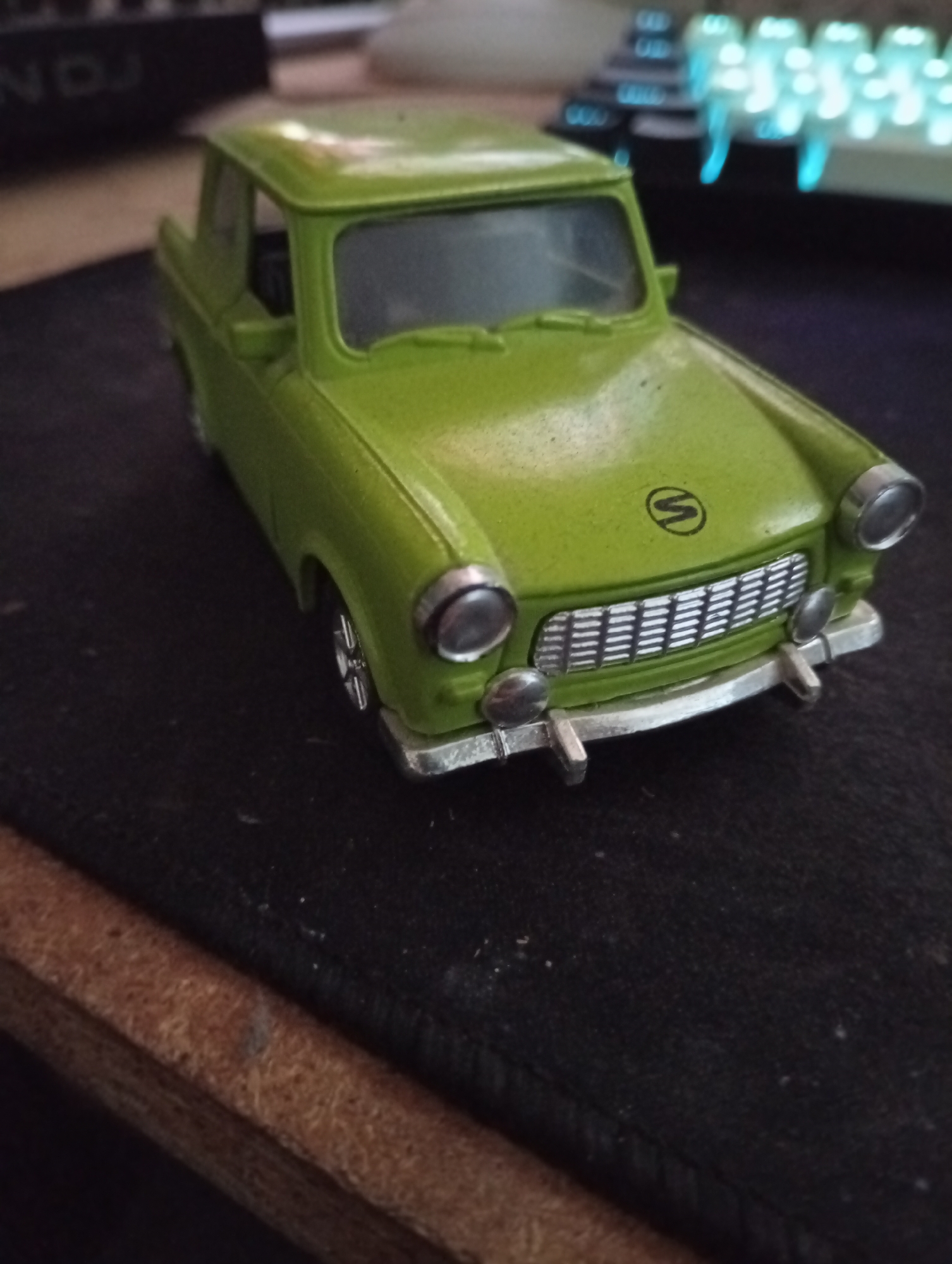 The image shows a close-up, high-angle shot of a green toy car, possibly a model of a Trabant, placed on a dark, textured surface. The car is positioned at a slight angle, with its front facing the viewer. The car's details, such as its headlights, grille, and emblem (a circular symbol with a stylized 'S' inside), are visible. The background is blurred, but suggests a desk environment with a keyboard emitting blue light and other electronic equipment. There is a wooden surface in the lower left corner. The image appears to be taken indoors. No specific location in the city of Wedel, Germany, can be determined from the visual information provided.