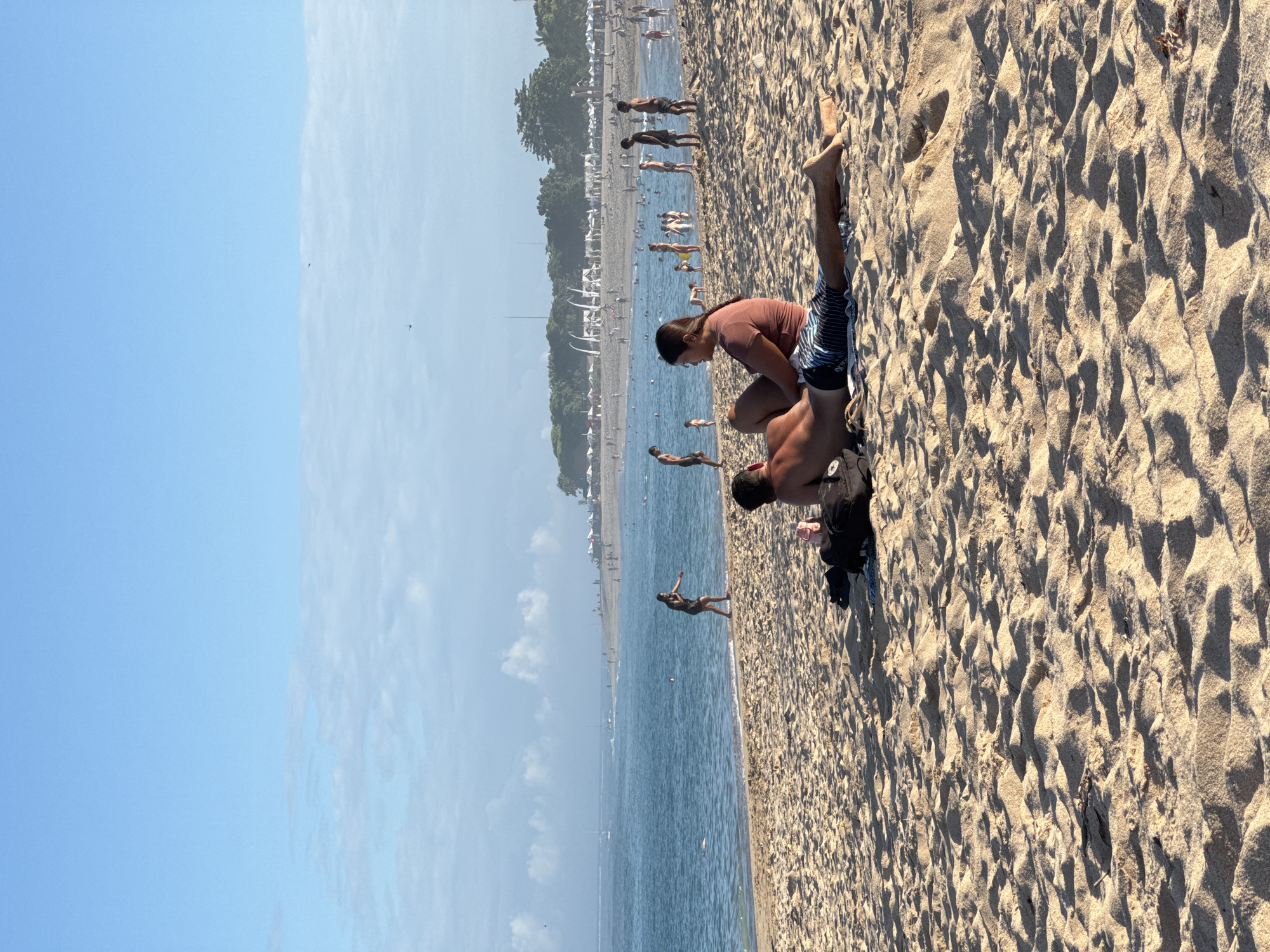 The image shows a sunny beach scene in Kuta Selatan, Indonesia. The sky is light blue with some clouds. The ocean is calm and stretches towards the horizon, meeting the beach. Several people are visible along the shoreline and in the water, including swimmers.
In the foreground, a couple is sitting on the sandy beach. A woman wearing a brown top and shorts is sitting next to a man wearing black shorts. They are positioned close to each other.
The beach is crowded with other people enjoying the sun and sea. White umbrellas and other structures are visible further along the beach. Trees and other greenery are visible inland. The sand is light-colored.