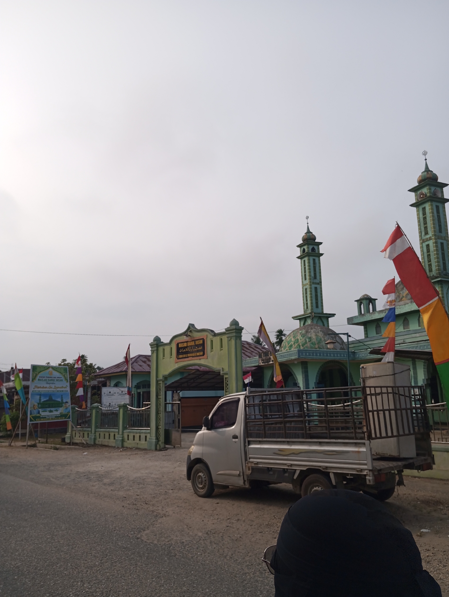 A medium shot shows the exterior of a mosque in Pematang Reba, Indonesia, with a light gray pickup truck parked in front of it. The mosque is painted a light green and features two tall minarets, a dome, and an arched entrance. Several flags, including red and white Indonesian flags, are visible in front of the mosque and on poles. A large banner with text and images is also displayed near the entrance. The truck has a metal cage in its bed. In the foreground, the head and shoulders of a person wearing a black hijab and glasses are visible. The ground is a mix of asphalt and dirt. The sky is overcast.