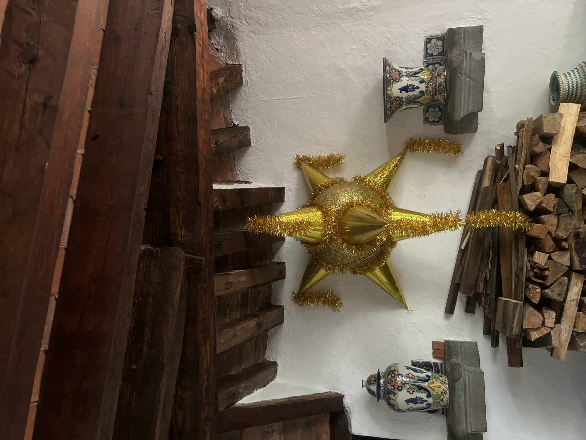 The image shows an interior setting in Mexico City, Mexico. A large, golden, star-shaped piñata hangs on a white wall. To the left is a dark brown wooden staircase. On the right, stacked firewood is mounted near two decorative ceramic vases affixed to grey stone. A woven basket sits above the firewood. The wall has a textured, stucco-like finish.