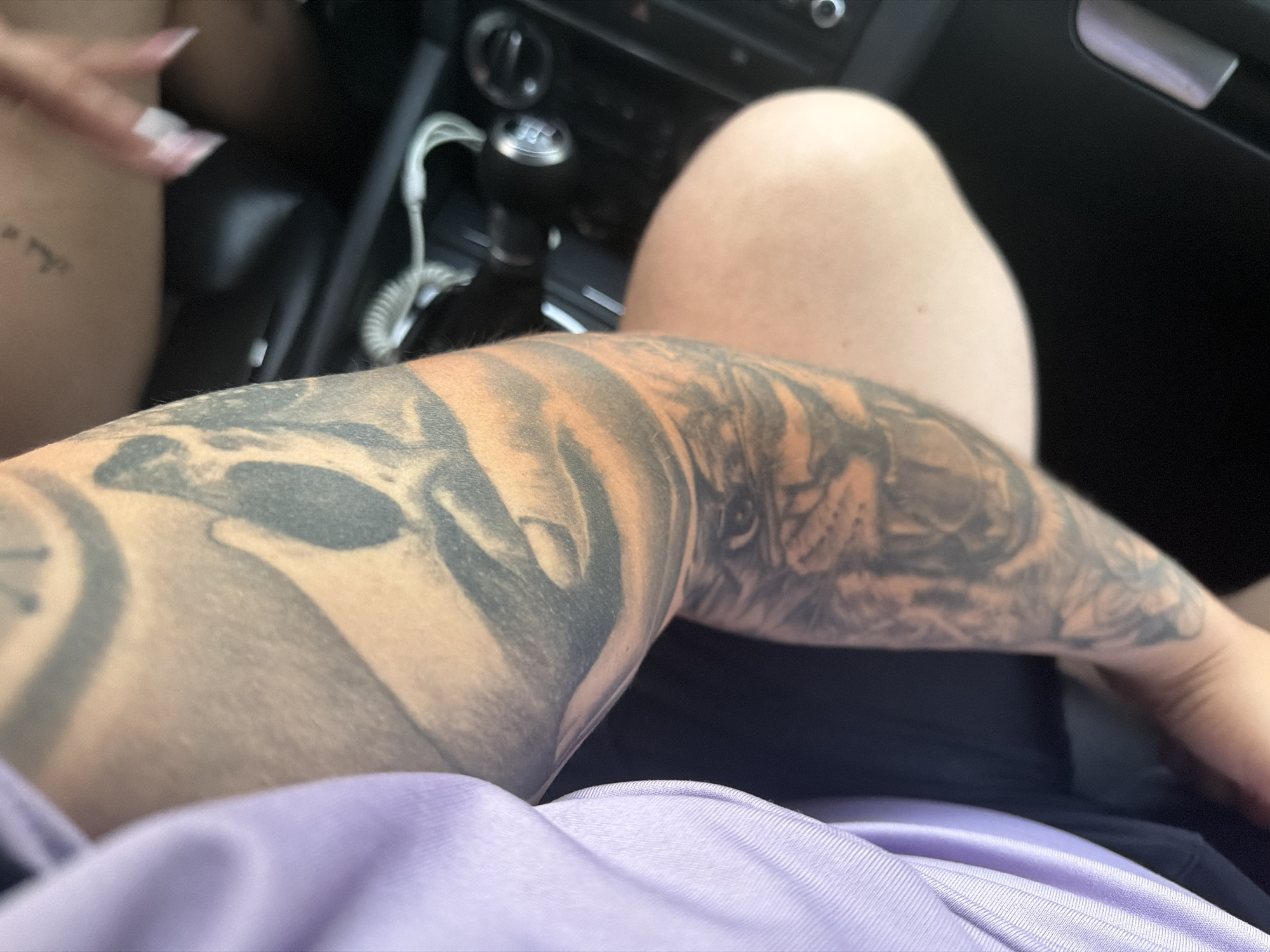The image captures the interior of a car, likely in Shumen, Bulgaria, during the daytime. The perspective is from a low angle, showing the center console and parts of two individuals.
In the immediate foreground, a person's arm, extensively tattooed with detailed imagery including a lion or tiger-like face and other patterns, stretches across the frame. The arm is partially covered by light purple or lavender fabric, likely clothing.
To the left, a portion of another person's bare leg or thigh is visible, adorned with a small, indistinct text tattoo that appears to be in a foreign script, possibly Hebrew, with characters resembling "וְאֵ֣י" at its start. This individual also displays light-colored, elongated fingernails.
The central focus of the car's interior is a manual gear stick, clearly marked with "R, 1, 2, 3, 4, 5, 6," indicating a six-speed transmission. Adjacent to it, sections of the dark-toned dashboard are visible, featuring rotary controls and air vents. A white coiled cable, possibly for charging or audio, is connected near the gear stick. A silver-colored door handle can be seen on the right side of the frame. The scene suggests a moment of rest or travel inside the vehicle.