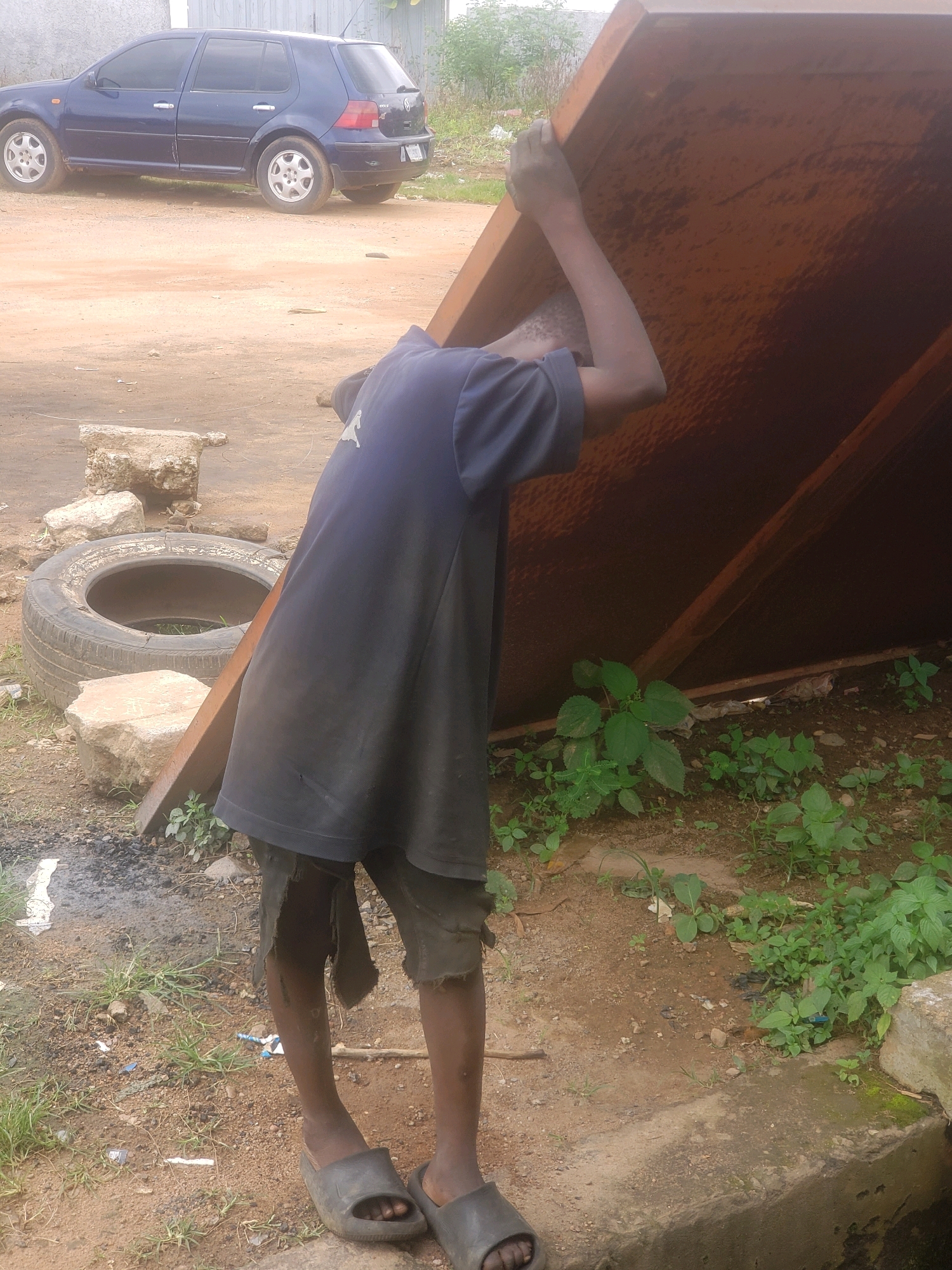 A young boy, wearing a dark blue t-shirt and ripped black shorts, is pictured in Sabon Tasha, Nigeria. He is lifting a large, thick, reddish-brown, flat, rectangular object, possibly a piece of metal sheeting, with his hands. The object is angled upwards and away from him. He is barefoot, wearing dark open-toed sandals. The ground is dirt and sand, with some patches of green weeds and scattered rocks. In the background, a dark blue four-door sedan is parked on a dirt road. A deflated black tire is visible on the ground to the boy's left, with a broken concrete block nearby. The lighting suggests it is daytime.