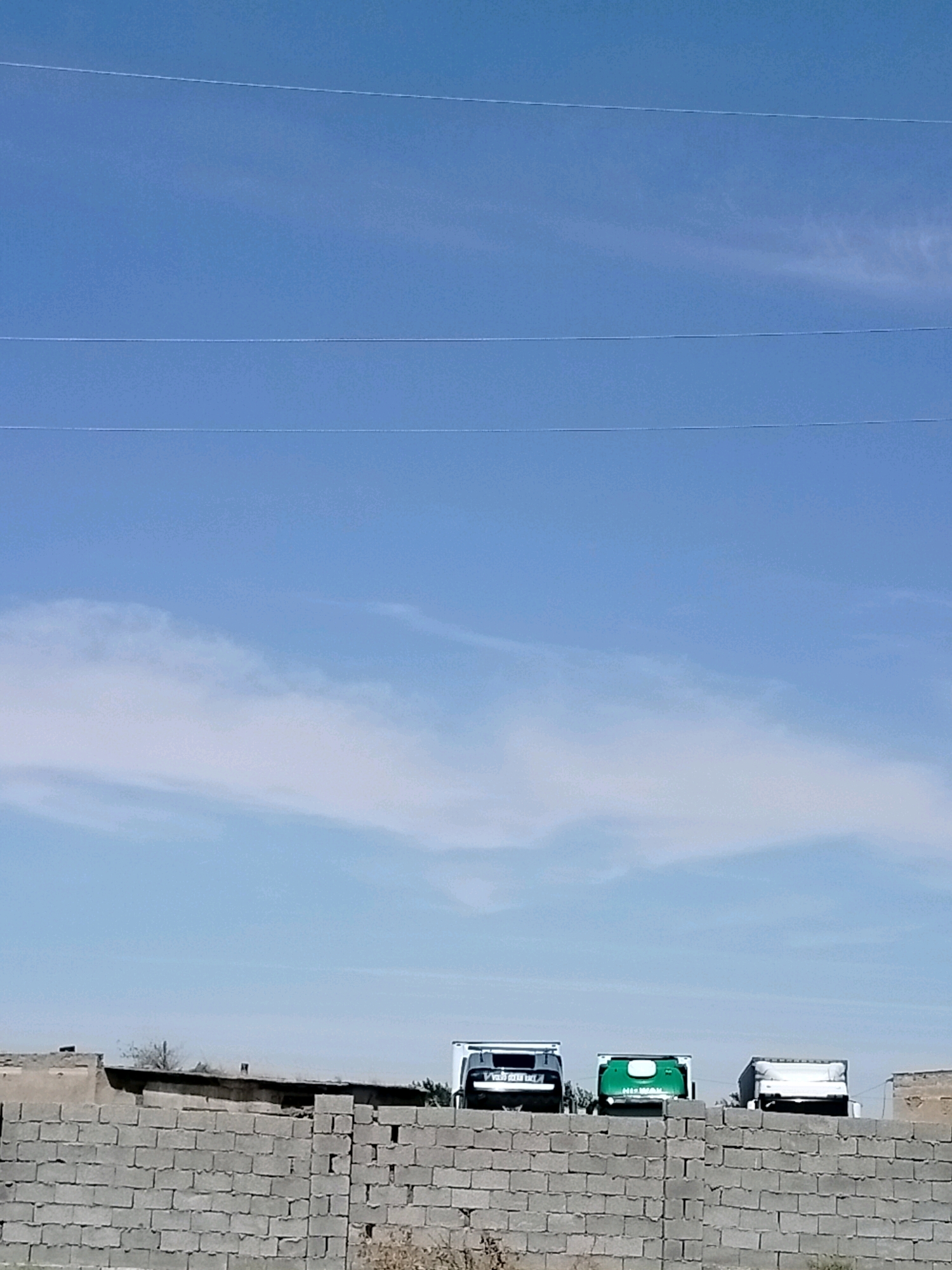 The image shows a bright blue sky with three thin, parallel white lines stretching horizontally across it, likely power lines. Below the sky, there is a band of white clouds. The lower part of the image features a scene with a brick wall in the foreground. Behind the wall, three trucks are parked in a row. The leftmost truck is white with black accents. The middle truck is green with white lettering on its rear. The rightmost truck is white with some black elements. The trucks are positioned on flat ground, with a few sparse trees and what appear to be low-lying buildings or structures visible behind them. The overall scene suggests an outdoor, possibly industrial or logistical area in or near the city of Көлкент, Kazakhstan, under a clear sky.