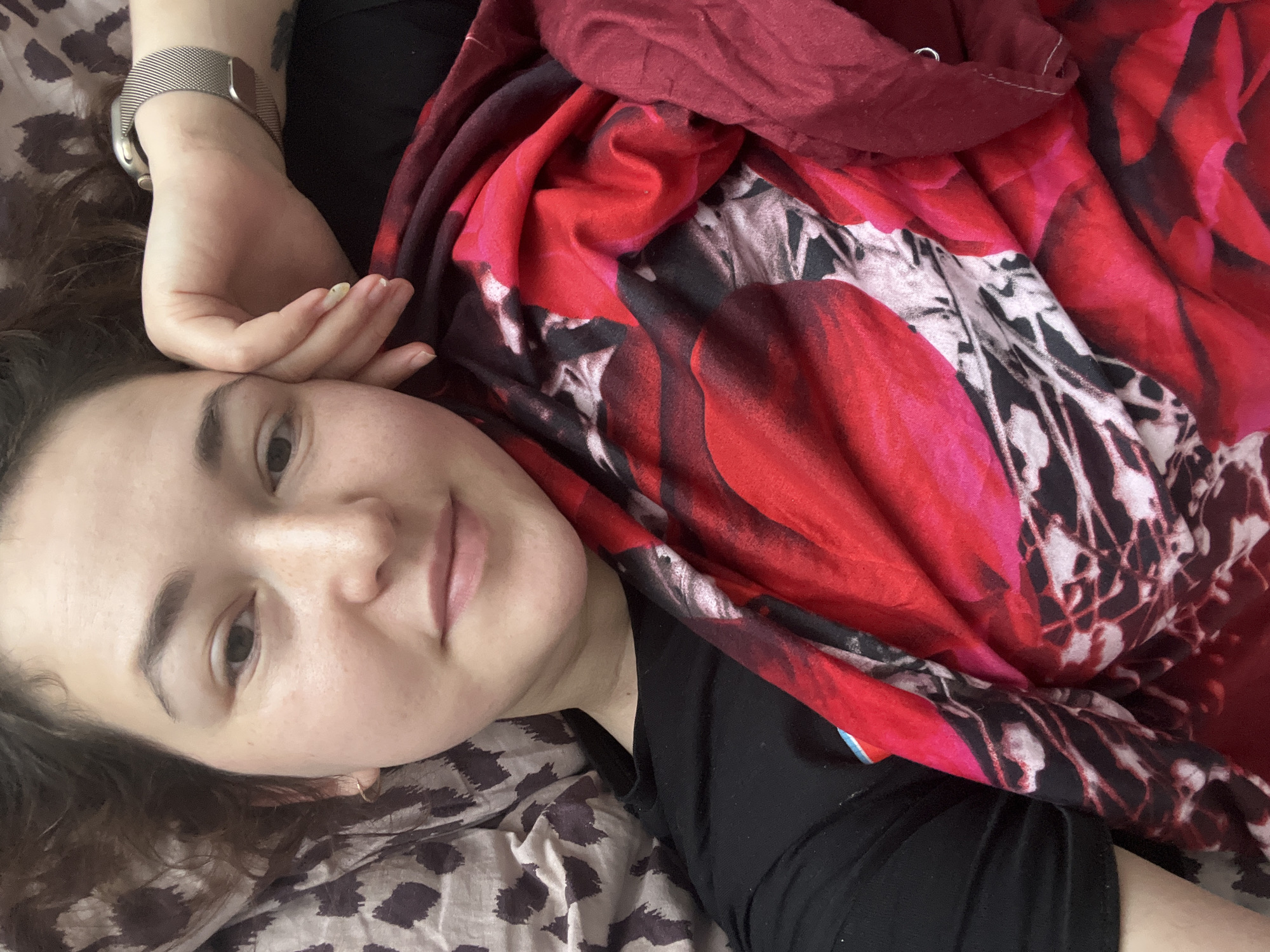 A selfie of a woman lying in bed, covered by a red and black floral patterned blanket. She is resting her head on her hand, with her left arm visible with a silver watch. The background consists of a pillow with a brown and white leopard print pattern. The woman has brown hair and fair skin with freckles. Her eyes are looking towards the camera. The image appears to have been taken indoors, likely in a bedroom. There is no information in the image to suggest a location in Chelyabinsk, Russia.