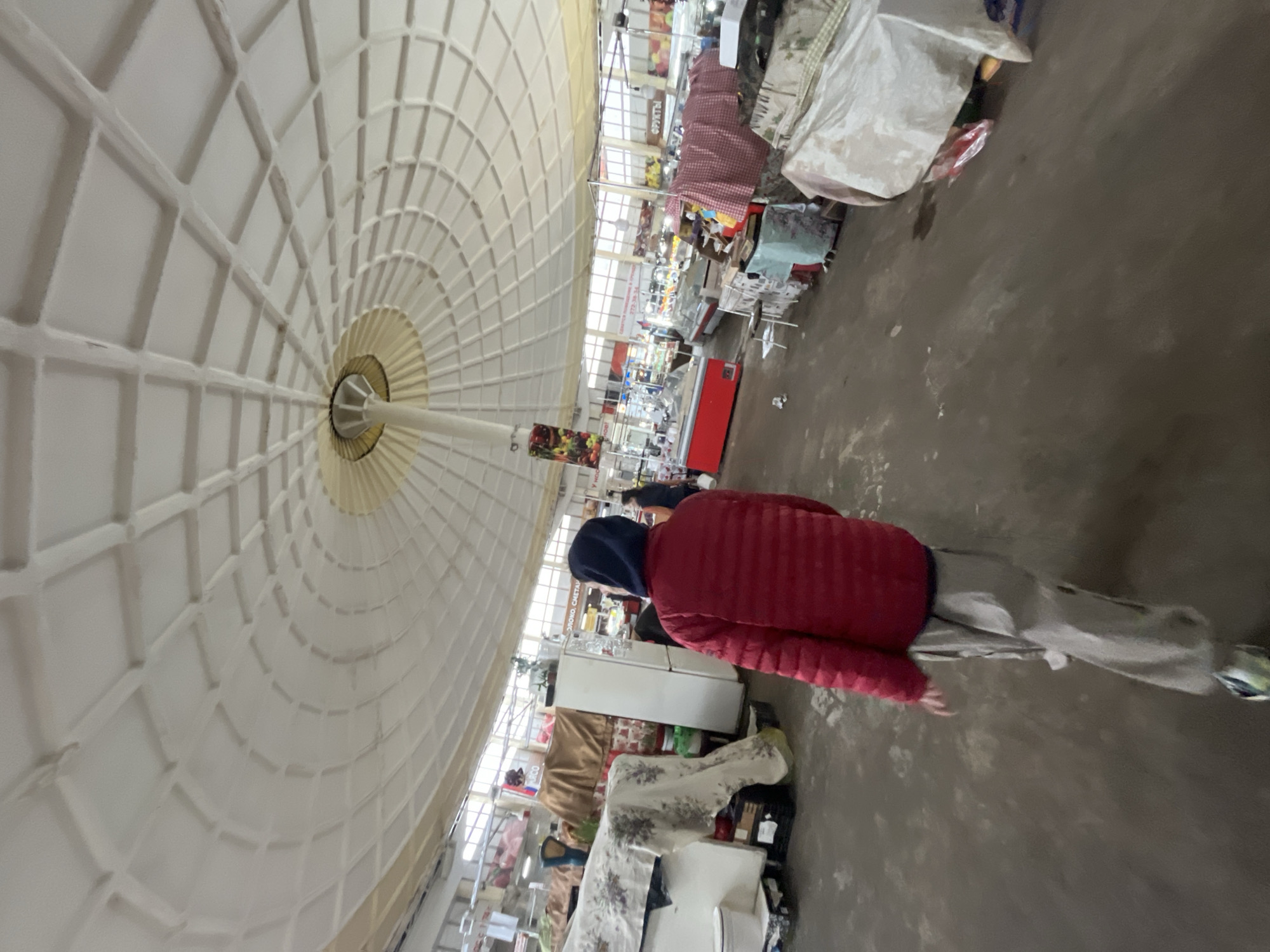 The image shows the interior of what appears to be a market, possibly in Unknown, Unknown, given the Cyrillic script visible on a sign. The roof is high and dome-shaped, with a complex grid pattern radiating from a central point.
A person wearing a red jacket and a blue hood is walking through the market. Stalls or tables display various goods, including fabrics and possibly food items. There are also refrigerators. The floor is a simple concrete surface. The lighting is bright and diffused, suggesting the market has a large area of open ceiling to allow light inside.