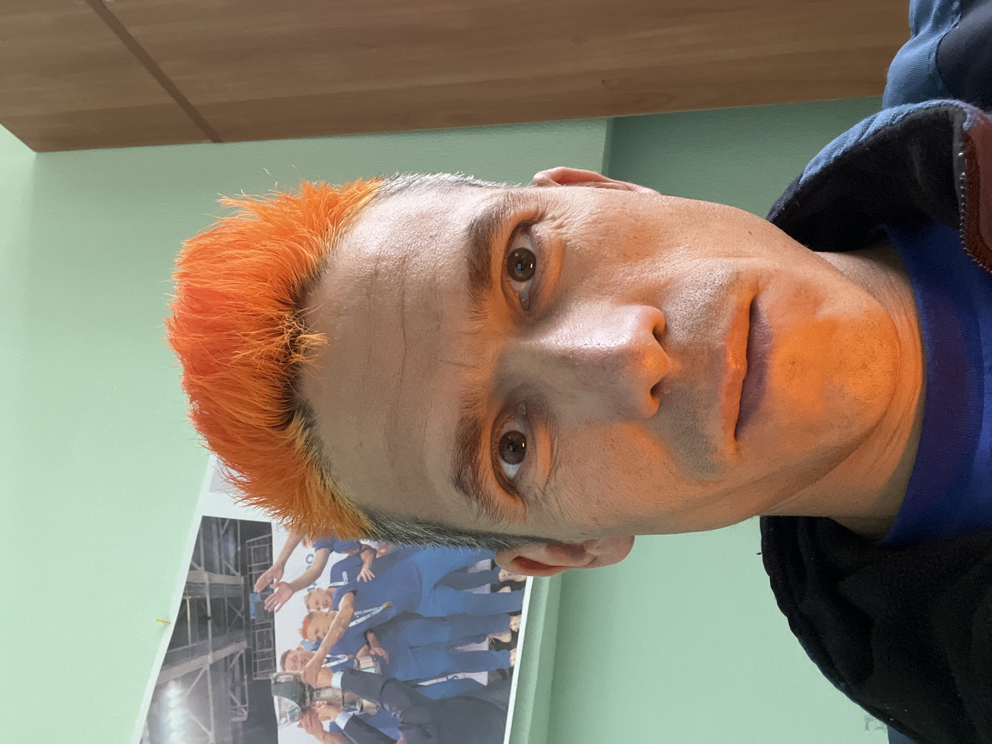 A close-up, rotated headshot of a middle-aged man with light skin, brown eyes, and graying hair dyed orange on top. He faces the camera with a neutral expression. He wears a dark-colored jacket with a blue shirt underneath. Behind him, a poster shows a group of people in blue suits raising a trophy or award. The backdrop is a light green wall with a wooden ceiling in the upper portion. The image may have been taken in an interior space in Chaikovsky, Russia, but there is not enough information to verify this.