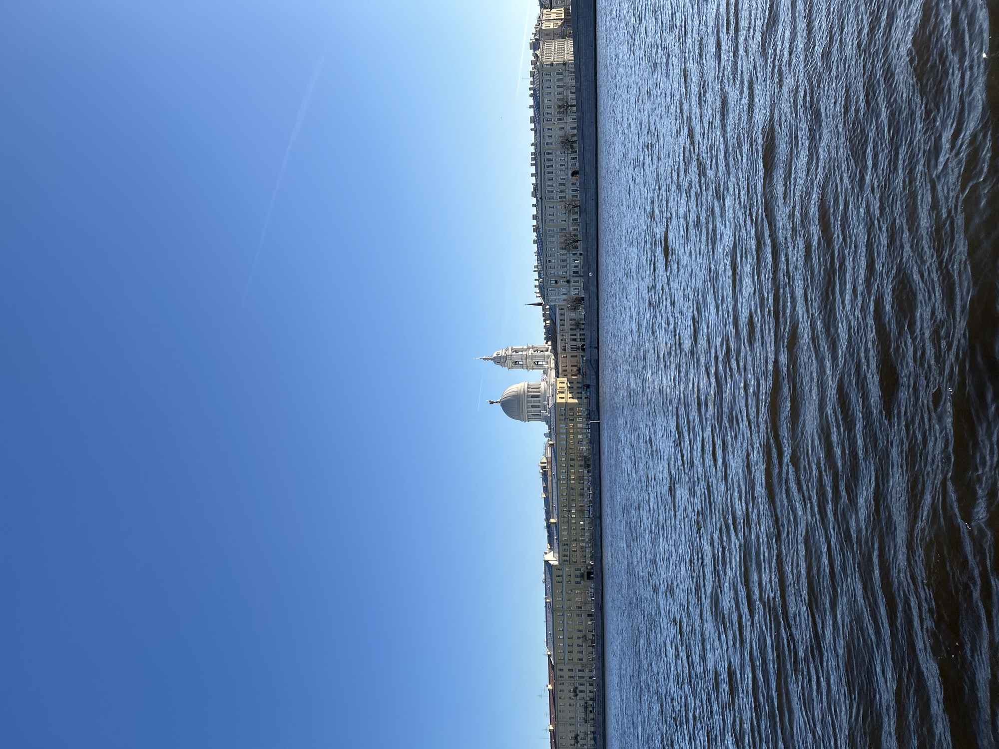 This panoramic image presents a serene urban landscape, likely Saint Petersburg, Russia, viewed across a wide body of water under a bright, clear sky. The composition is divided into three horizontal layers: a vast blue sky at the top, a city skyline in the middle, and a wide river at the bottom.
The sky is a vibrant, cloudless blue, indicating a sunny day. A faint, thin white contrail streaks diagonally across the upper left section, suggesting a distant aircraft.
Occupying the middle ground is the distinct skyline of a European city. A long stretch of elegant, multi-story buildings lines the horizon, characterized by classical architectural styles and numerous evenly spaced windows. These buildings appear light-colored, possibly beige or pale yellow, and are well-preserved. A prominent white edifice with a grand dome and an adjacent smaller dome or tower stands out slightly to the left of the center. This architectural feature strongly suggests a significant landmark in Saint Petersburg, such as Saint Isaac's Cathedral, confirming the provided location context.
The bottom portion of the image is filled by a wide expanse of dark blue water, which is heavily textured with countless small, parallel ripples and waves. These patterns suggest a gentle current or breeze, creating varying reflections of light across its surface. This body of water is characteristic of a major river or canal in Saint Petersburg, such as the Neva River.
The scene conveys a sense of peaceful grandeur. There are no people visible, and no specific activities are taking place, presenting a quiet, static view of the city. The time of day is clearly daylight, under optimal weather conditions. No visible text is present in the media file.