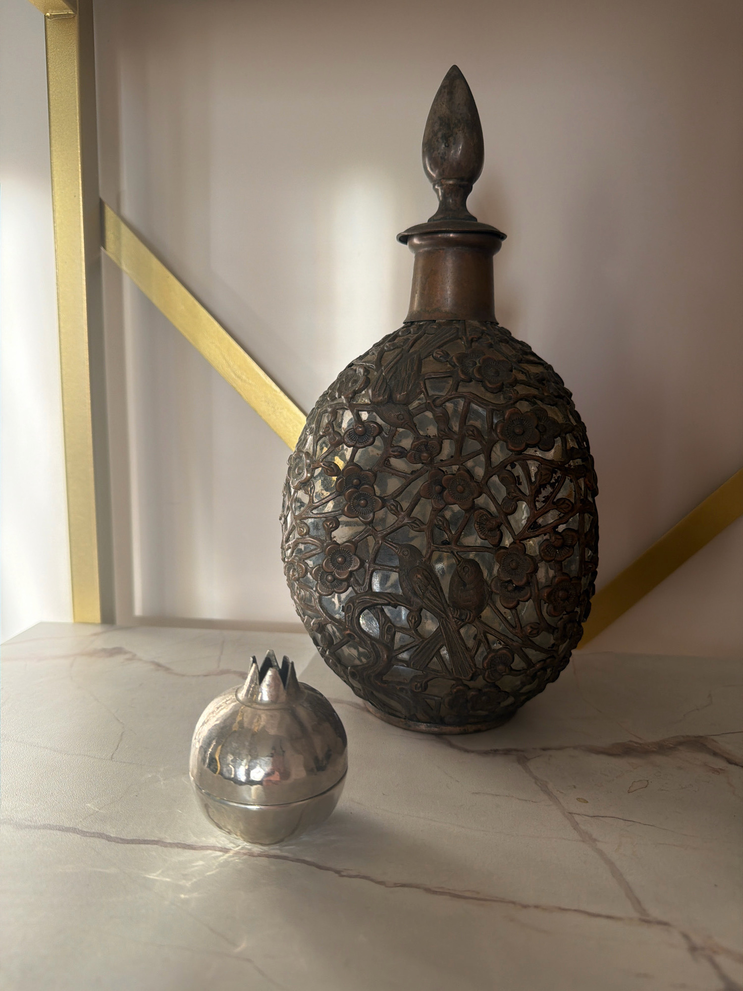 The image shows a decorative brass bottle with an ornate filigree design featuring birds and flowers, placed next to a small, silver pomegranate-shaped container. Both items are displayed on a marble-like surface. In the background, a modern shelf with gold accents is visible against a plain wall. The lighting creates shadows and highlights on the objects and surface, suggesting an indoor setting. There are no people present in the scene. The image does not provide any information that would place it in the city of Samara, Russia.